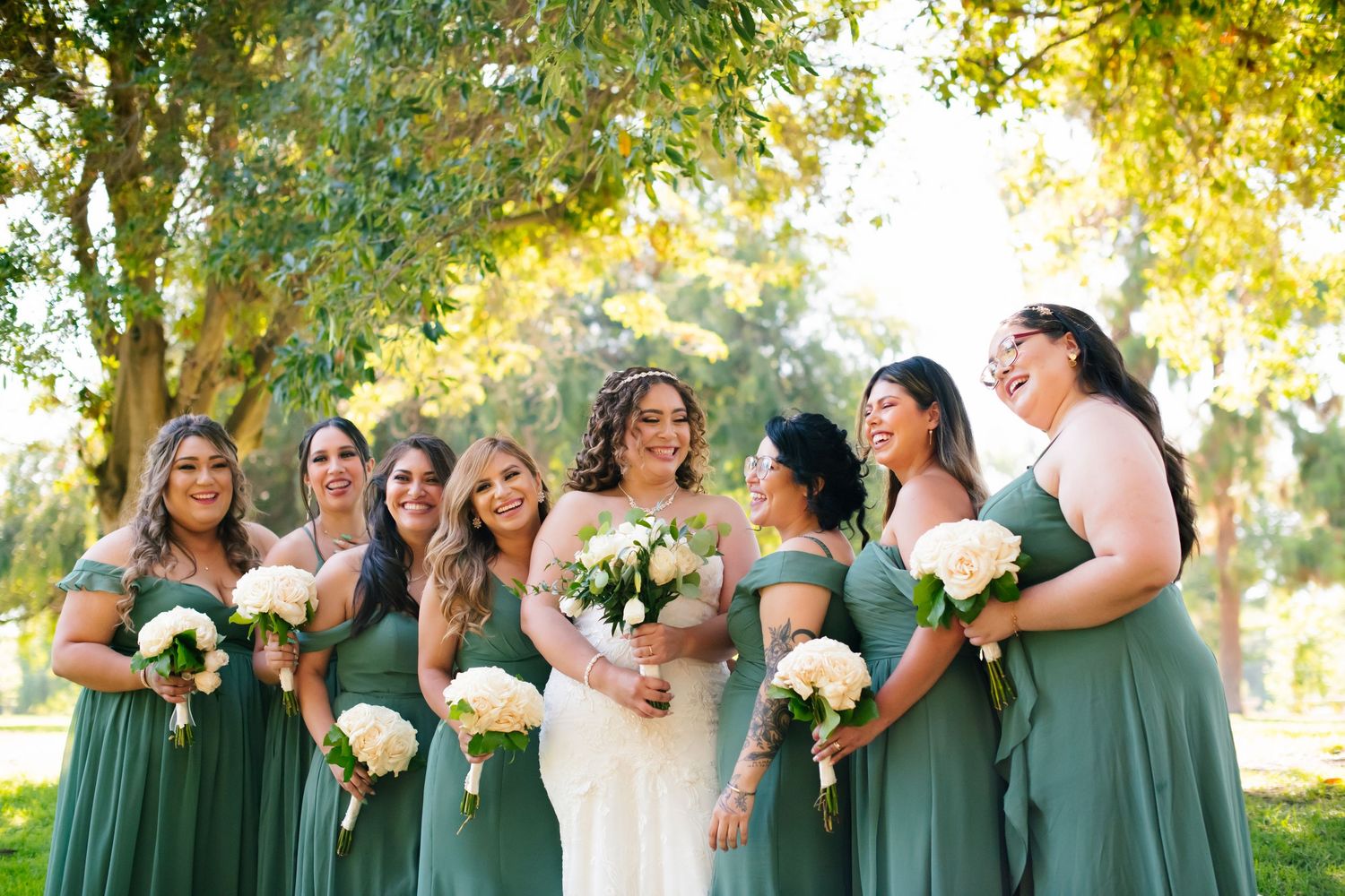 Bridesmaids in sage green gowns stand with bride for outdoor wedding photos.