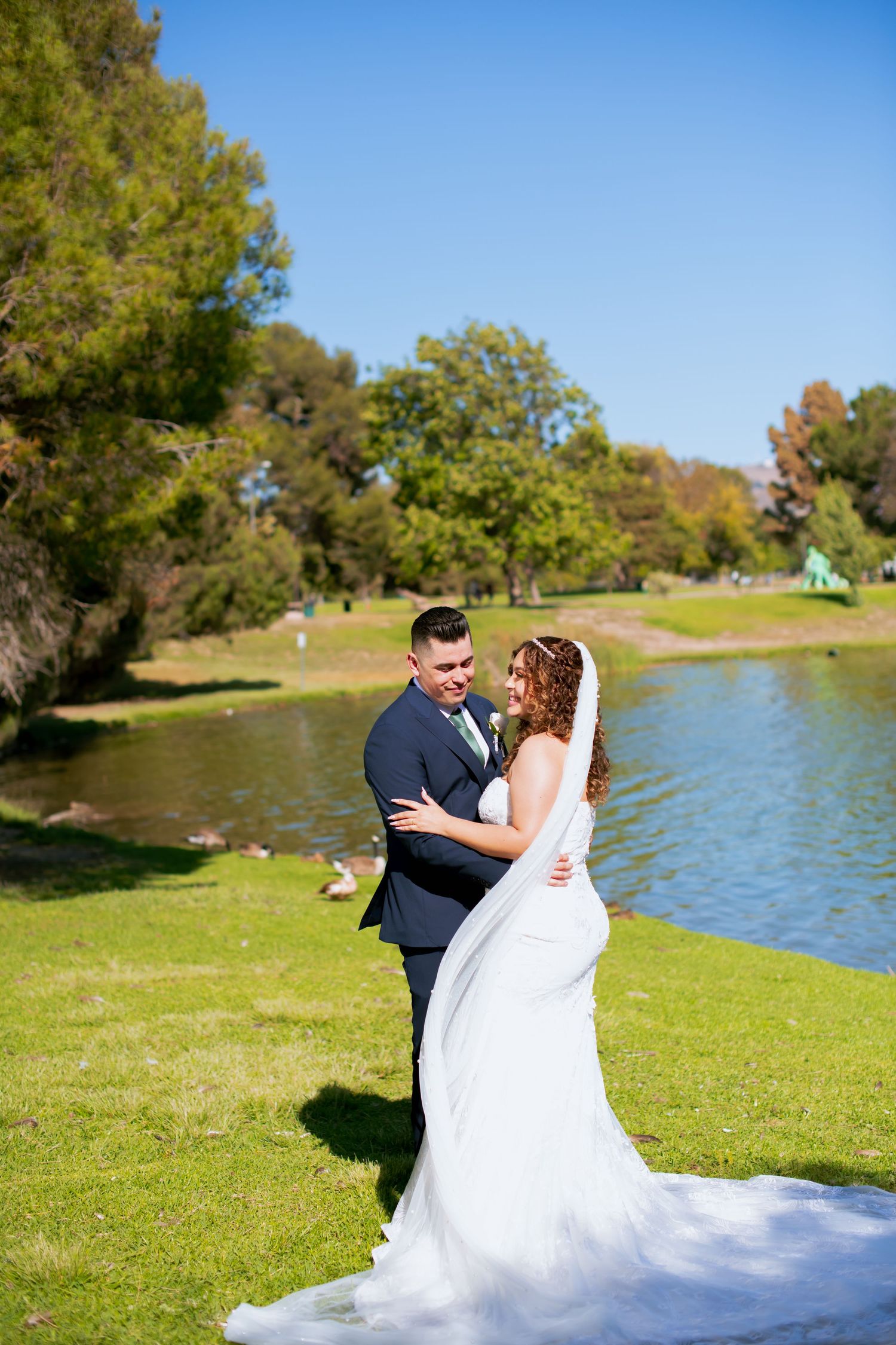 Wedding couple shares intimate moment on grassy lawn with lake and trees in background.