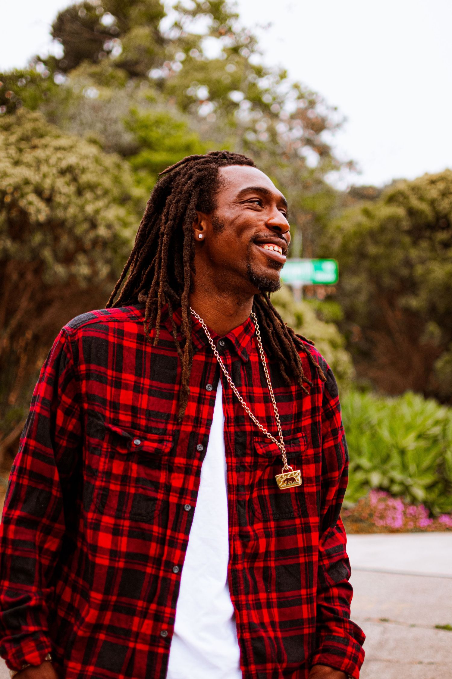 A person in red plaid flannel shirt smiling outdoors on a sunny day with lush greenery in the background.