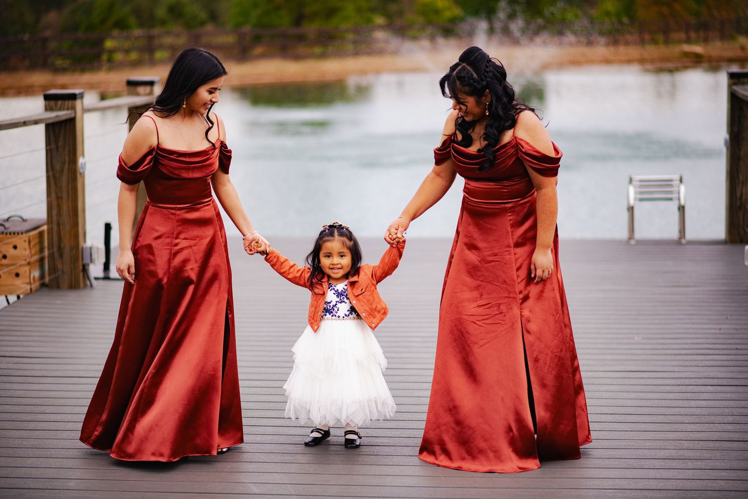 A group of people in rust-colored dresses walk along a wooden boardwalk near a lake.