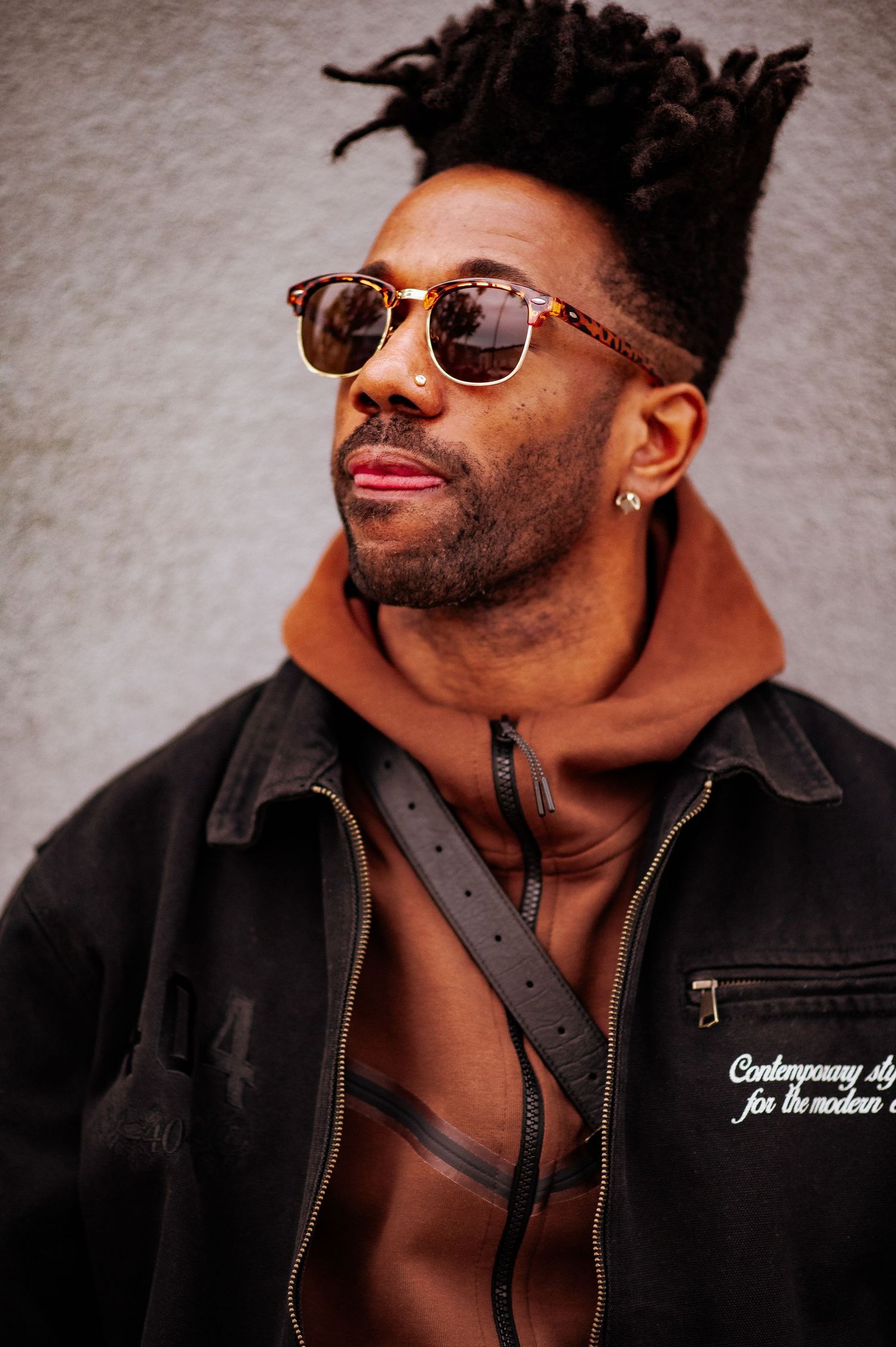 A stylish man in sunglasses and black jacket poses confidently against a gray backdrop in a fashion photoshoot series.