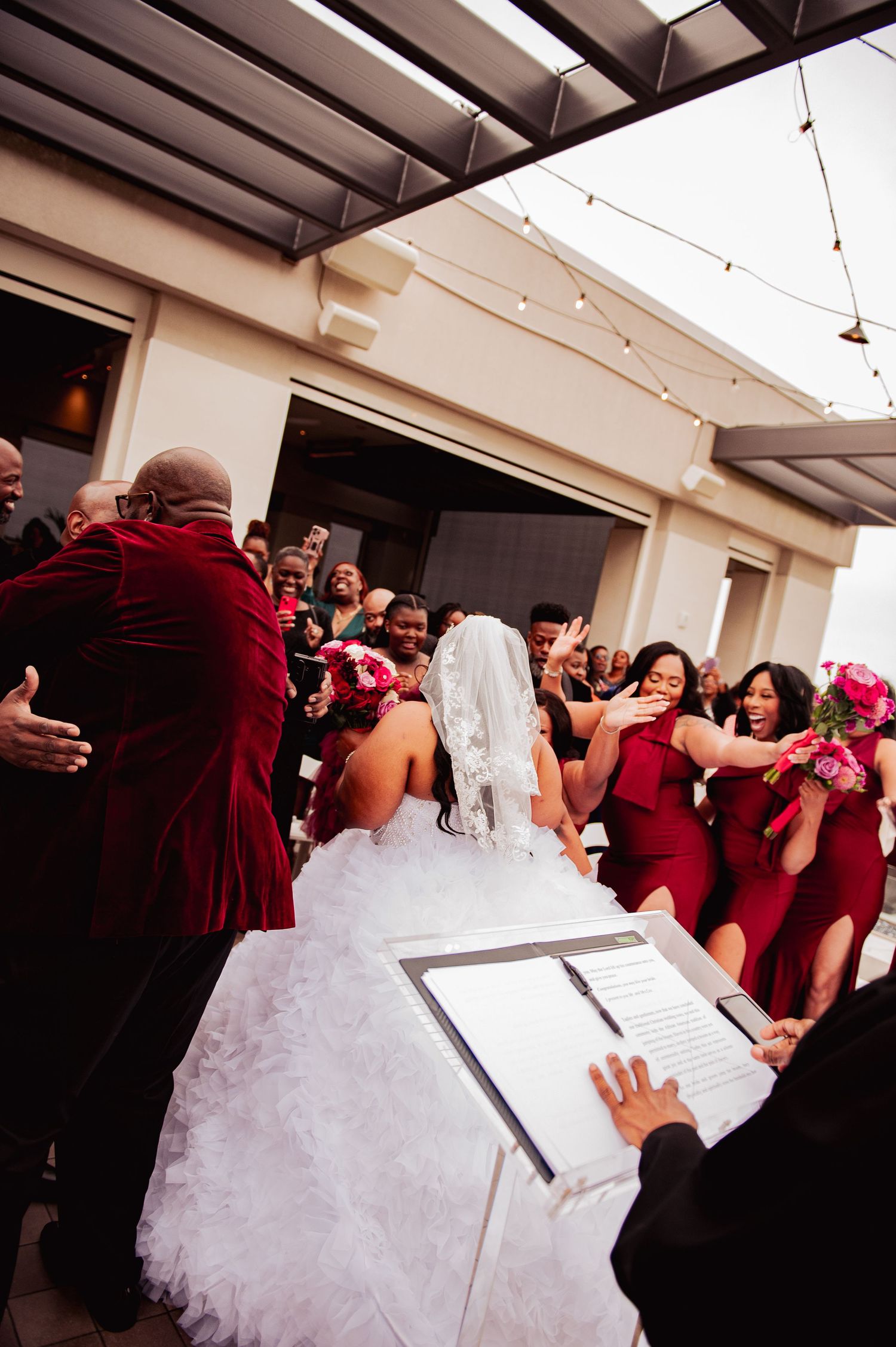 Wedding party in burgundy attire gathers around bride in white gown during outdoor ceremony signing moment.