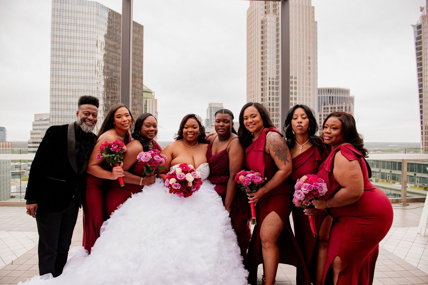 Wedding party in red dresses and black suits posed on urban rooftop with city skyline backdrop.