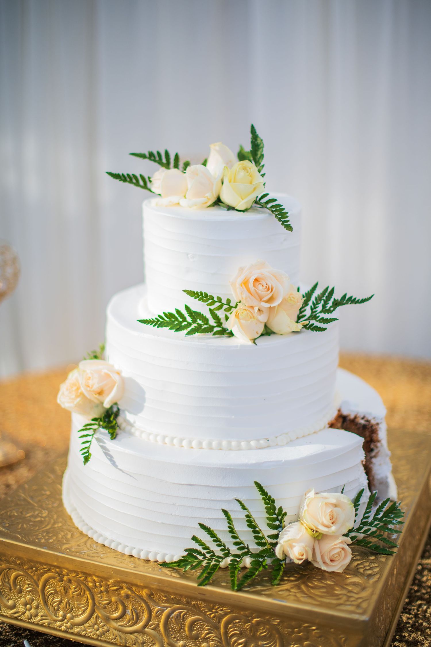 Elegant white wedding cake decorated with blush roses and ferns displayed on an ornate gold stand.