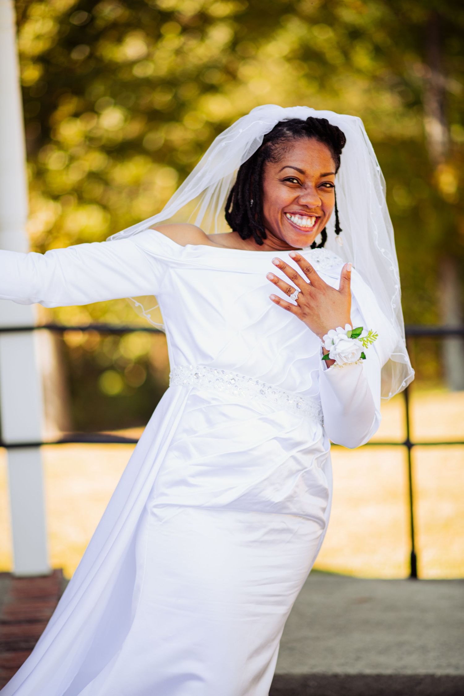 Bride in flowing white wedding dress poses playfully outdoors in natural sunlit setting.