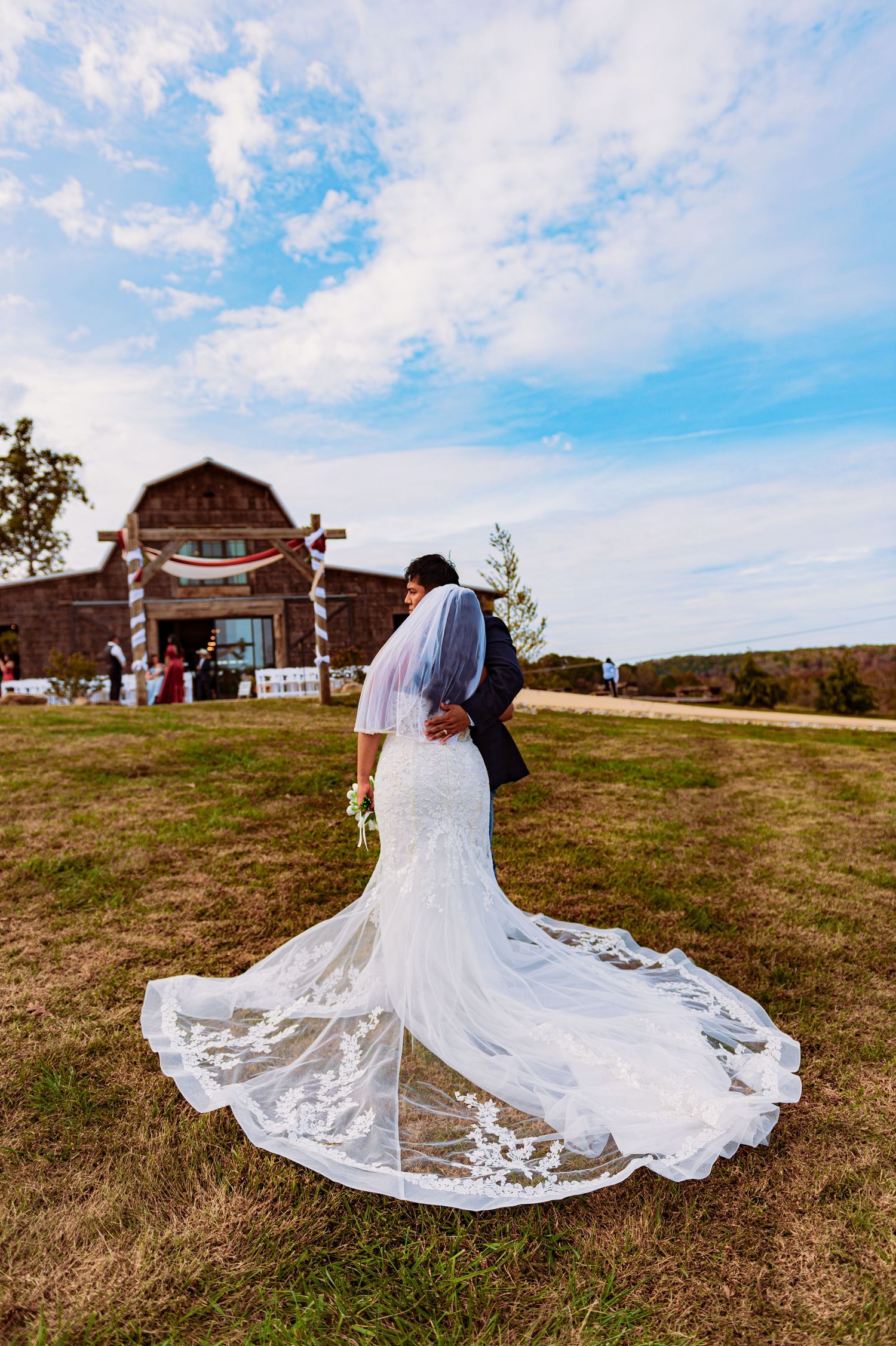 A flowing white wedding dress with intricate lace details spreads across a grassy hilltop with a rustic barn in the background.