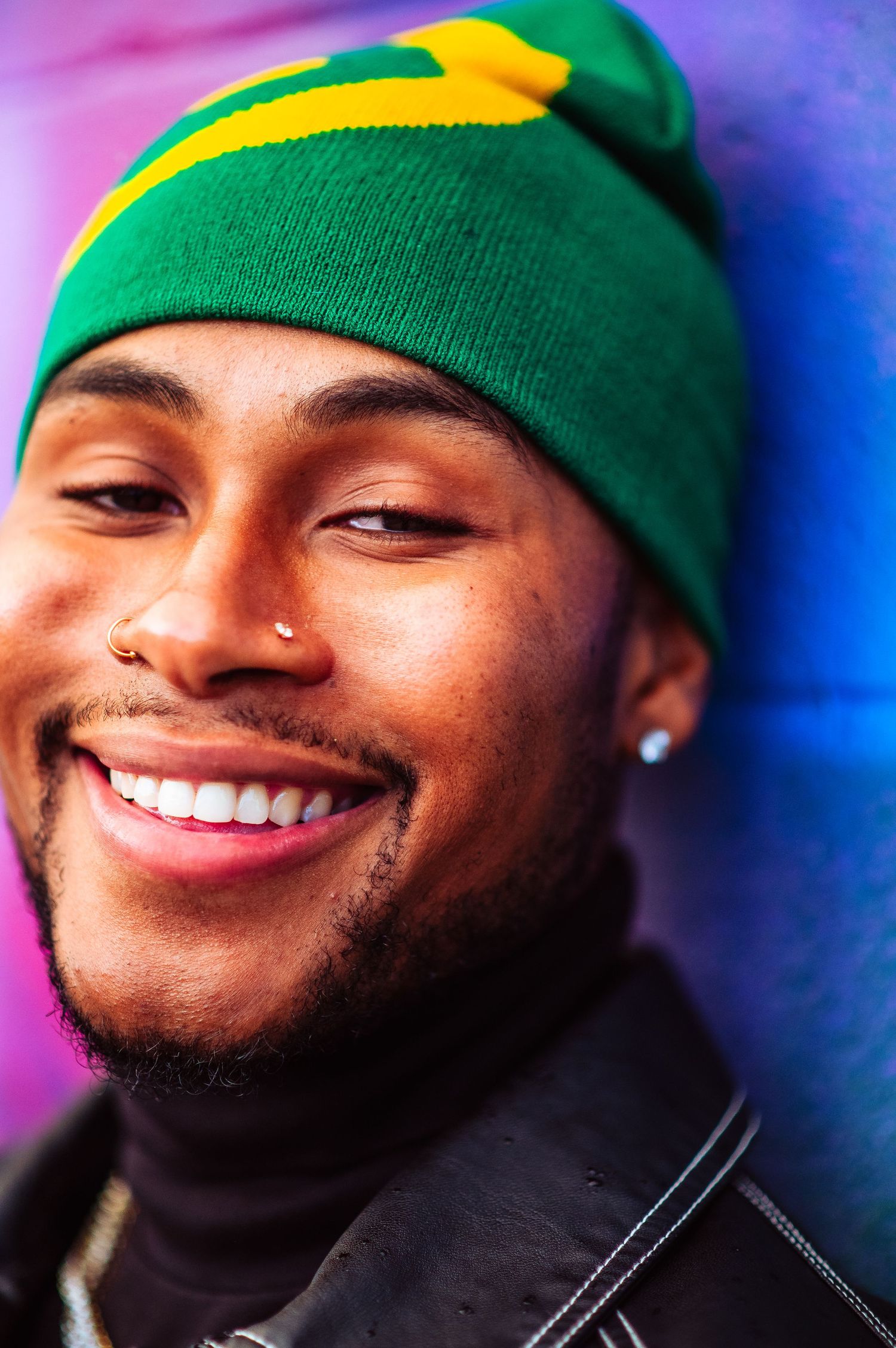 A bright smile radiates warmth against a colorful background, wearing a green and yellow beanie and black jacket.