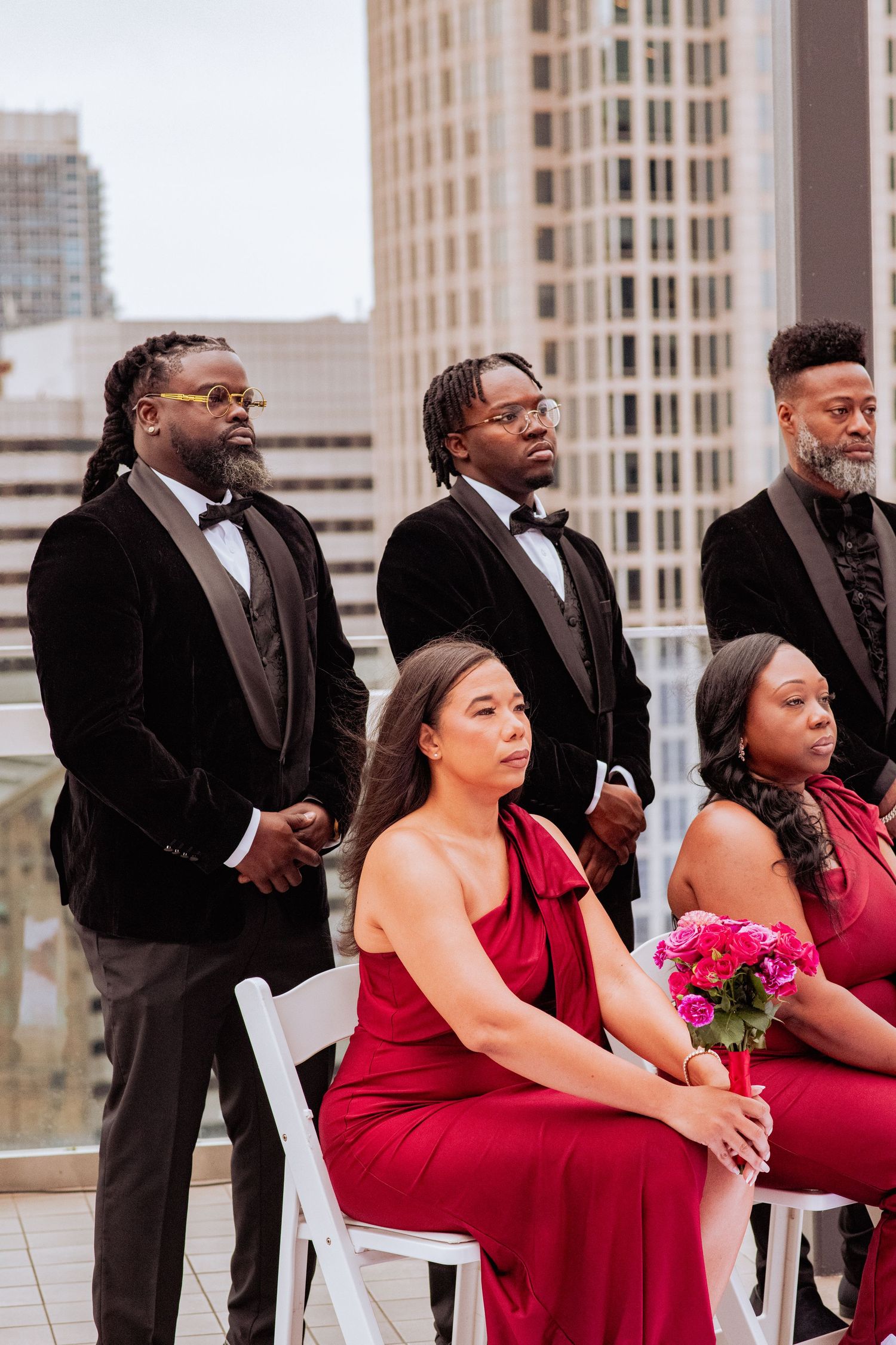 Wedding party members in burgundy dresses and black suits sit in white chairs during an outdoor ceremony.