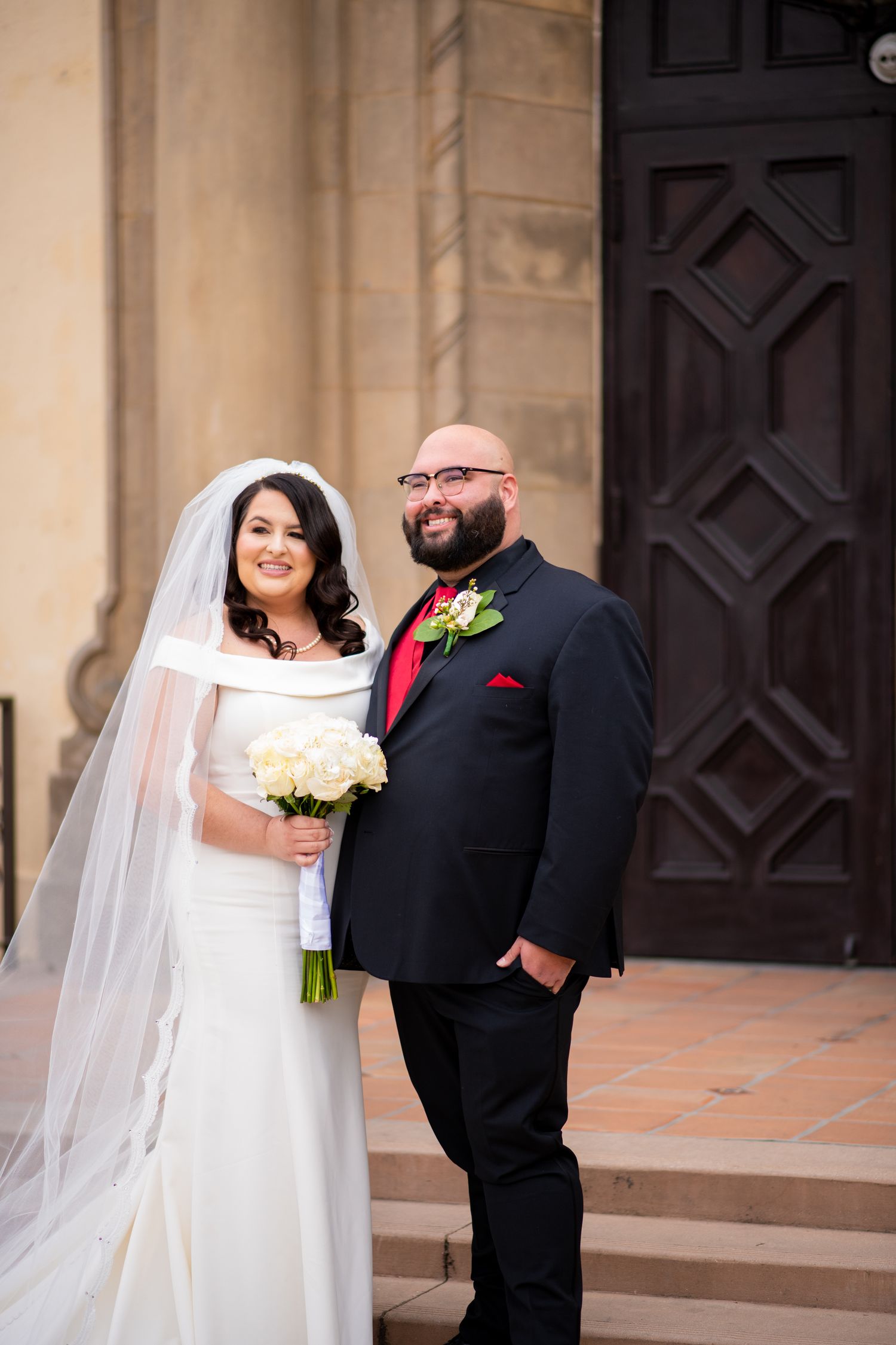 A wedding couple stands on stone steps in front of a dark wooden door, with the bride holding a white bouquet.
