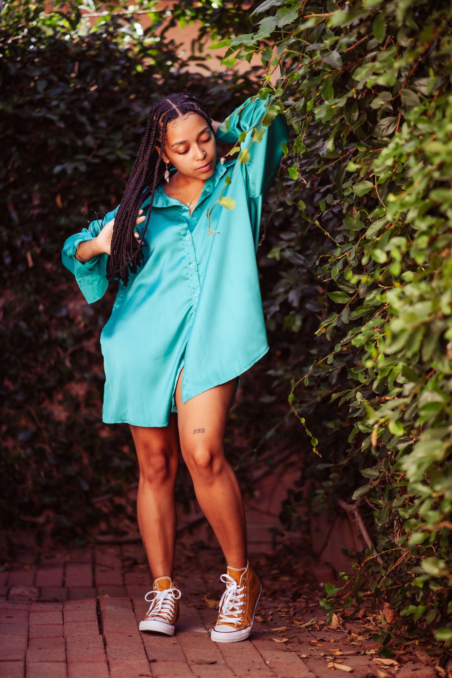 A person in a turquoise shirt and brown sneakers poses playfully against a backdrop of green foliage and brick pavement.