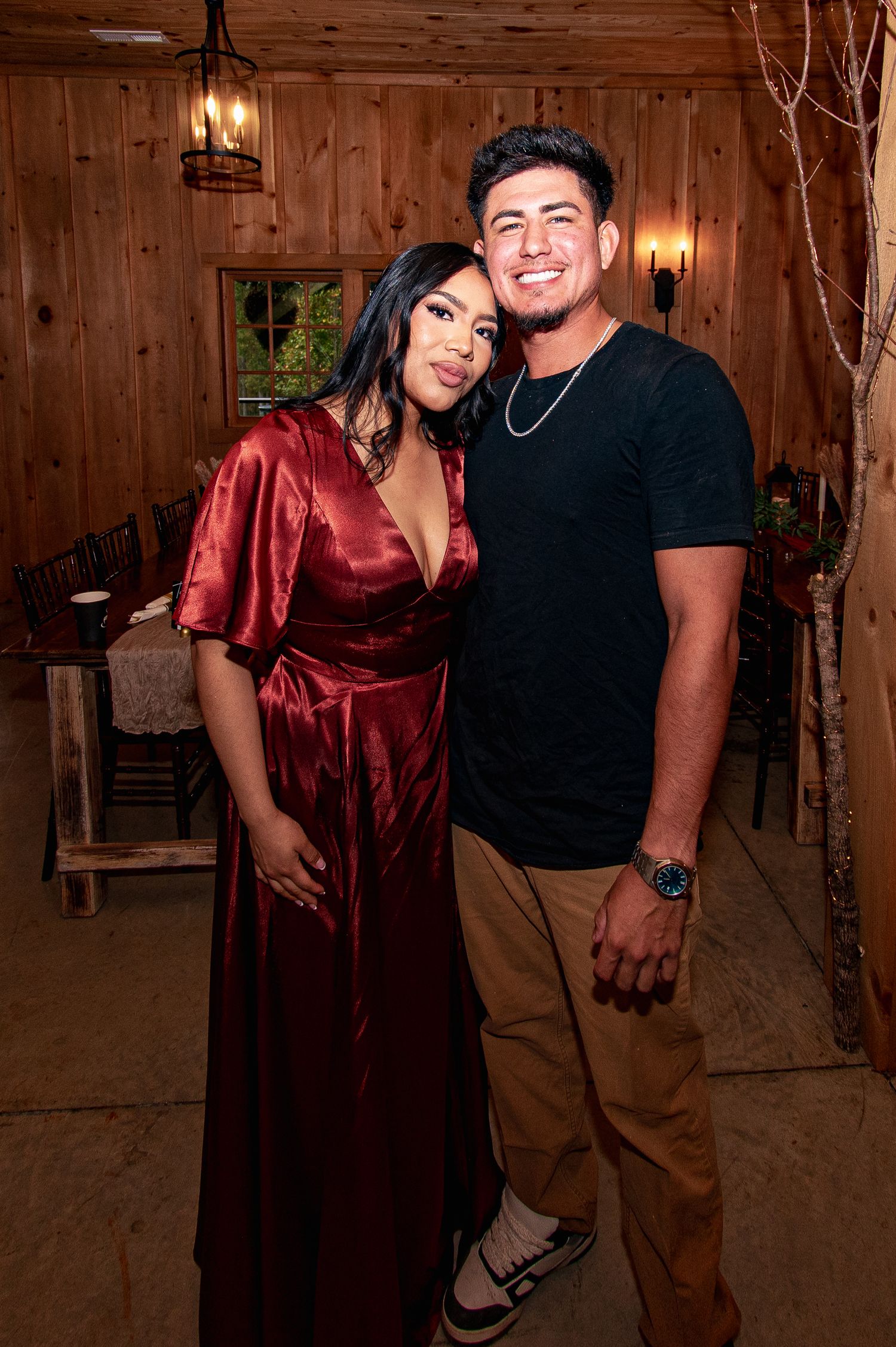 A couple in a rustic wooden interior poses playfully together, with one wearing a burgundy satin top and khaki pants.