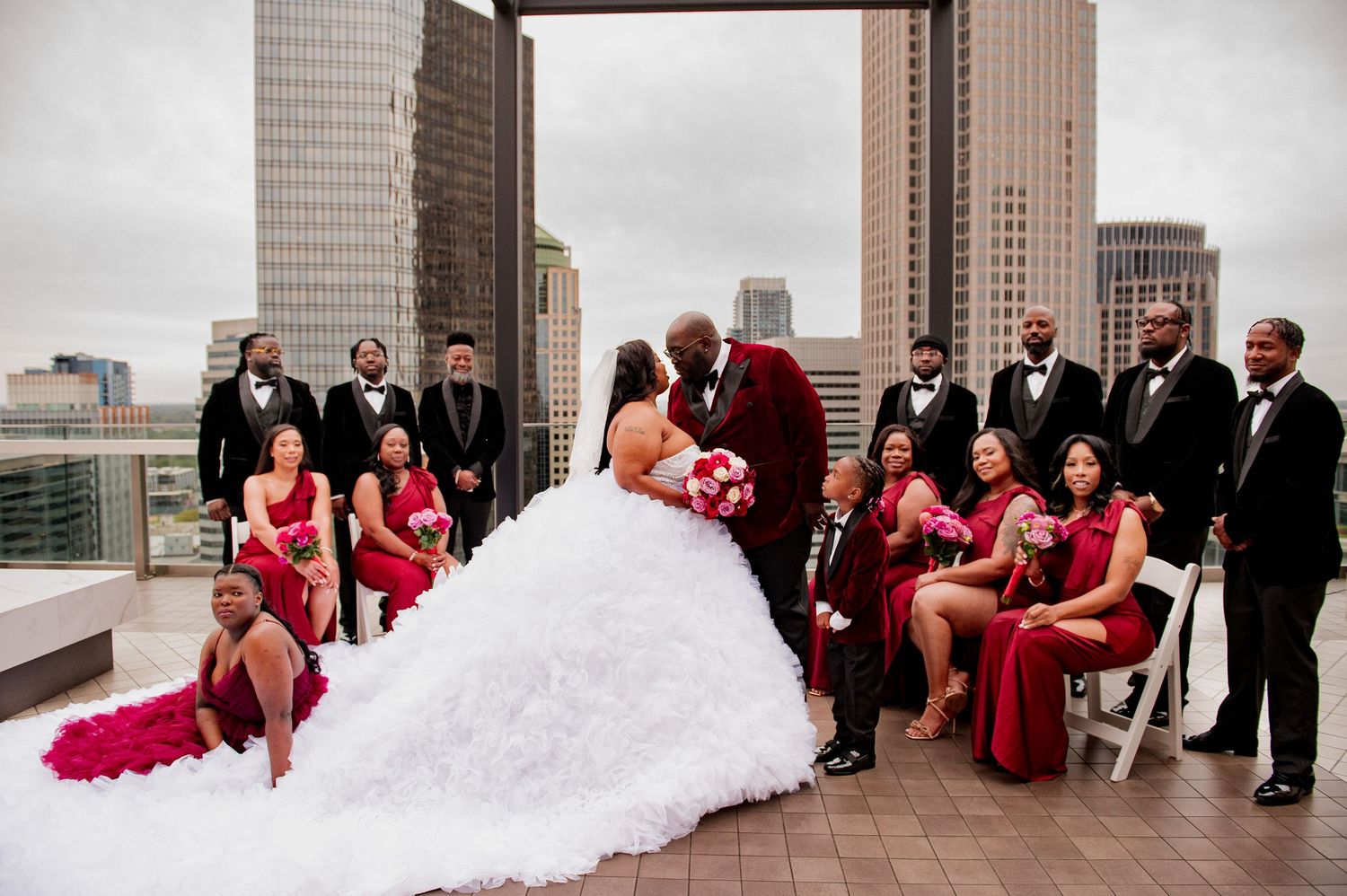 Wedding party in formal attire gathered on a rooftop terrace with bridesmaids in red dresses and groomsmen in black suits.