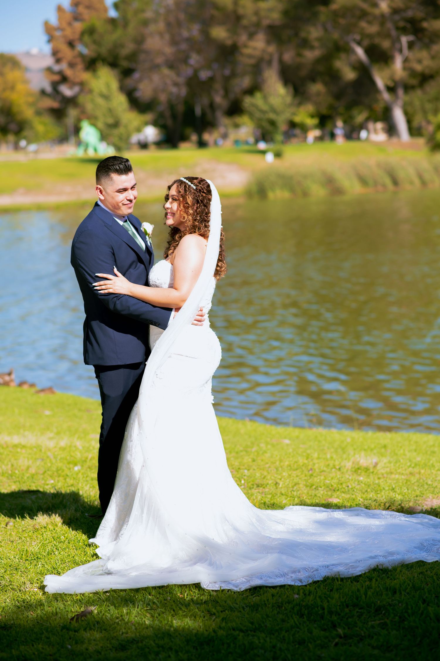 Romantic wedding couple embraces on grassy lawn beside lake during outdoor photoshoot.