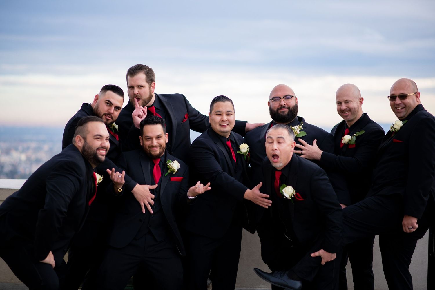 A group of groomsmen in black suits pose playfully for a series of candid wedding party photos outdoors.