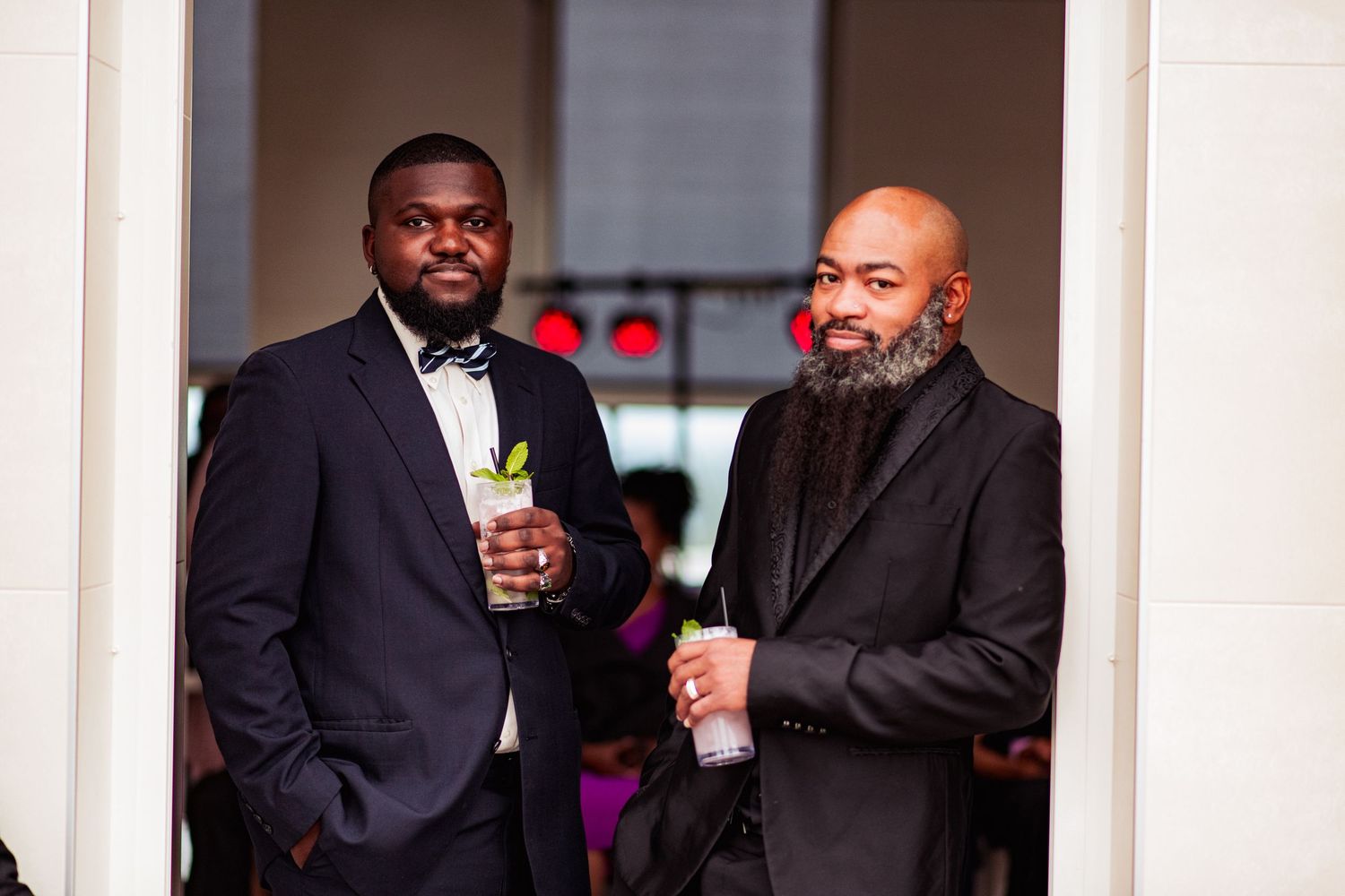 Two men in formal attire stand together at an event, one wearing a black suit and the other with a full beard.
