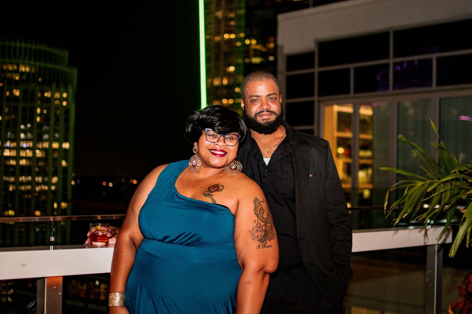 A couple poses together at night on a rooftop with city lights visible in the background.