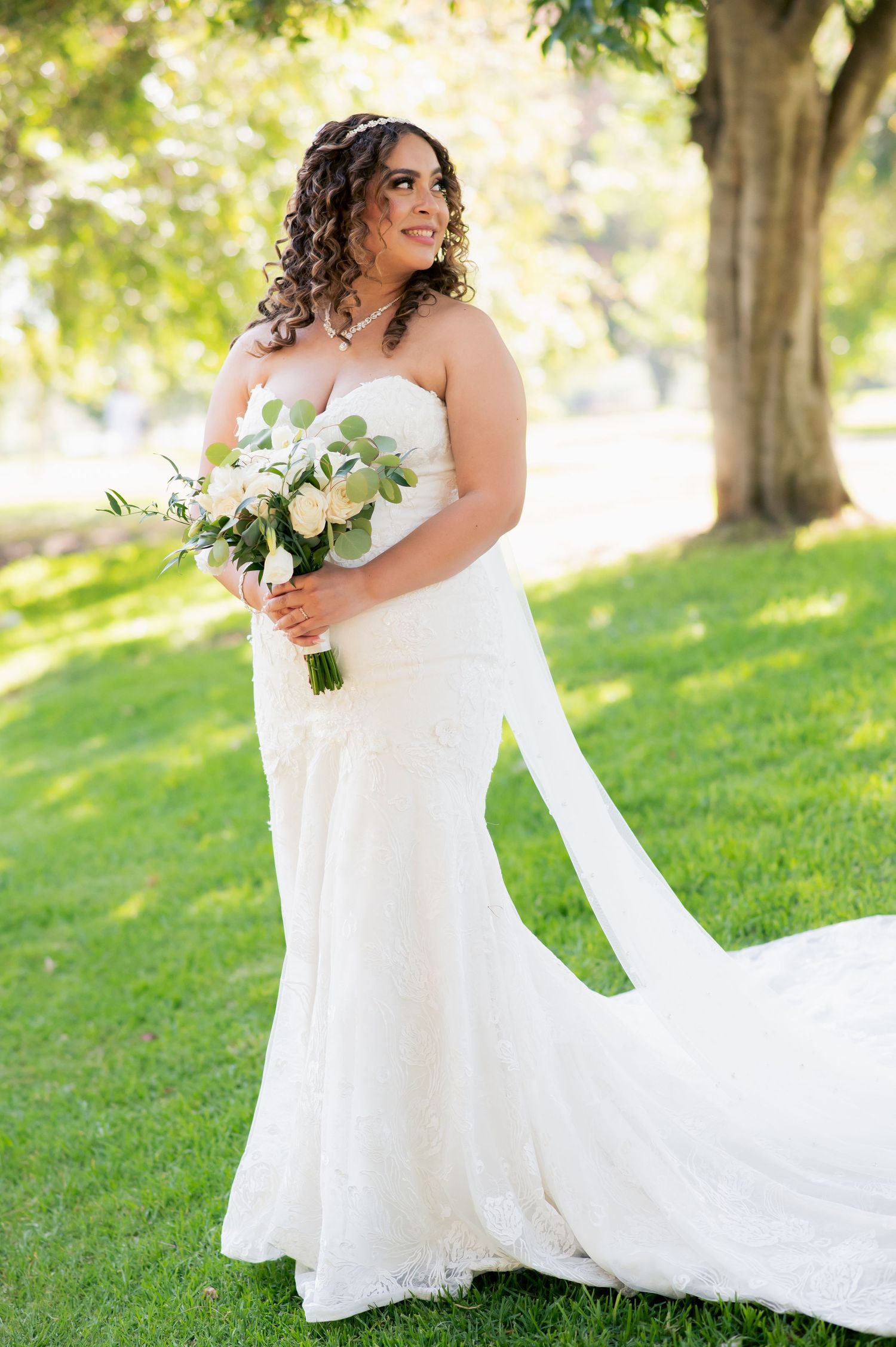 A flowing white wedding dress cascades across sun-dappled grass in an outdoor setting.