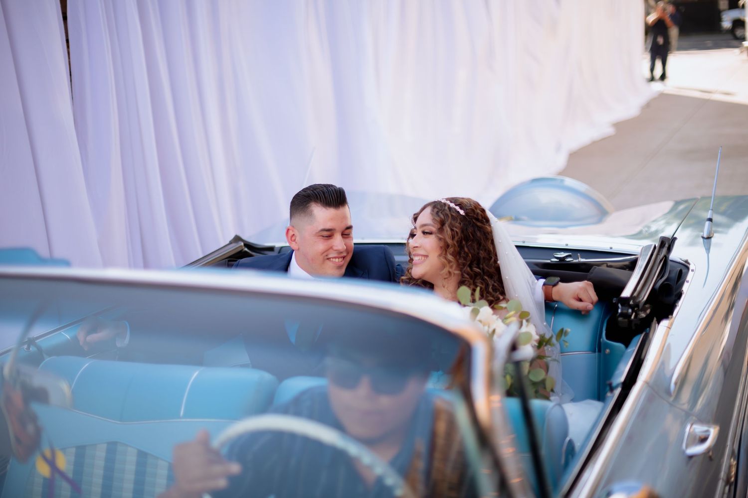 Wedding party members exit ceremony near vintage blue car with white draping in background.