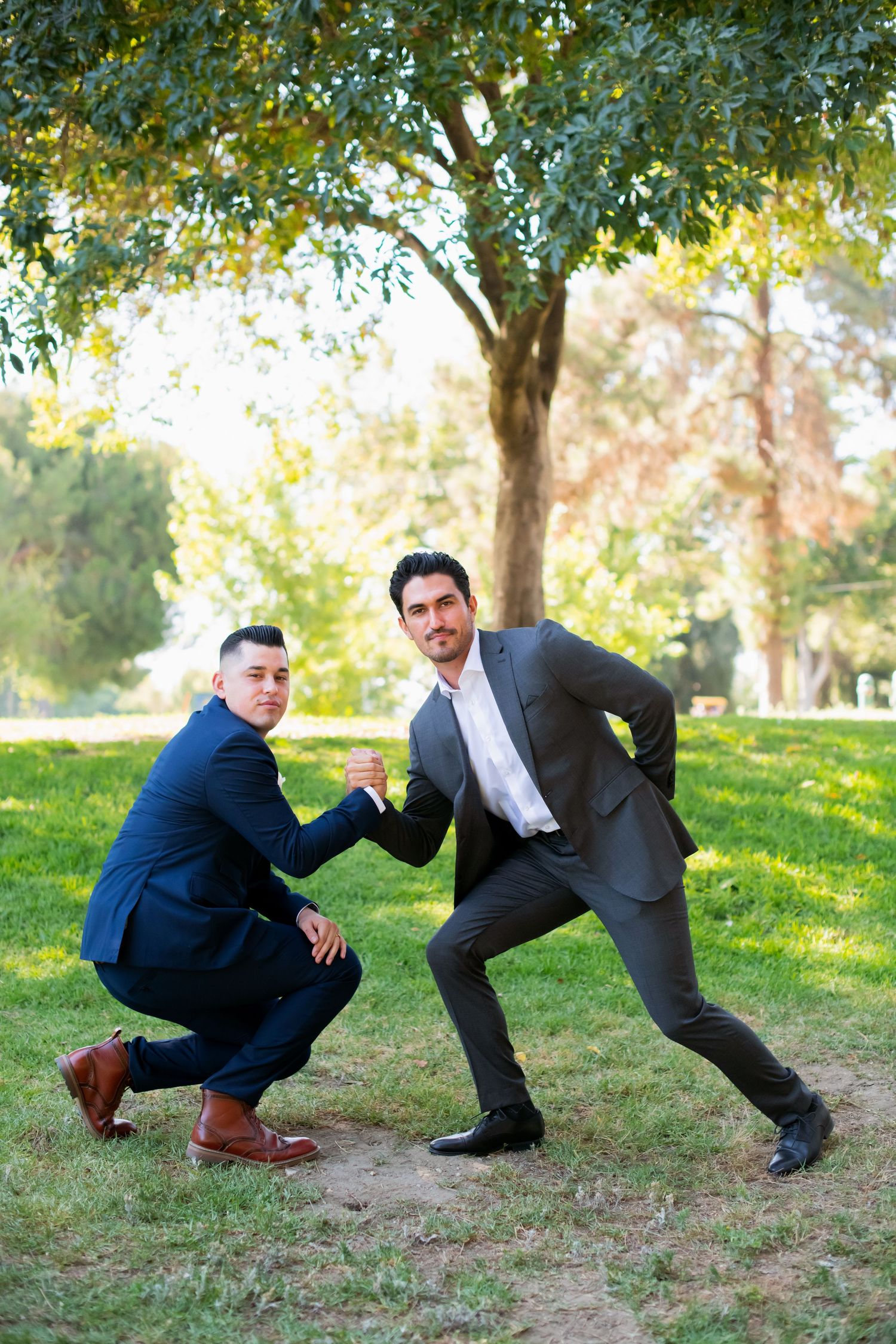 Two people playfully posing in crouching positions on grass under trees.
