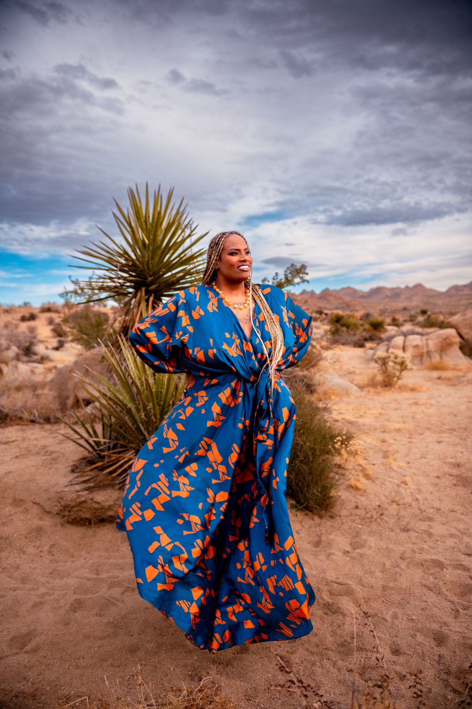 Person in flowing blue floral dress twirls gracefully in desert landscape with dramatic cloudy sky.