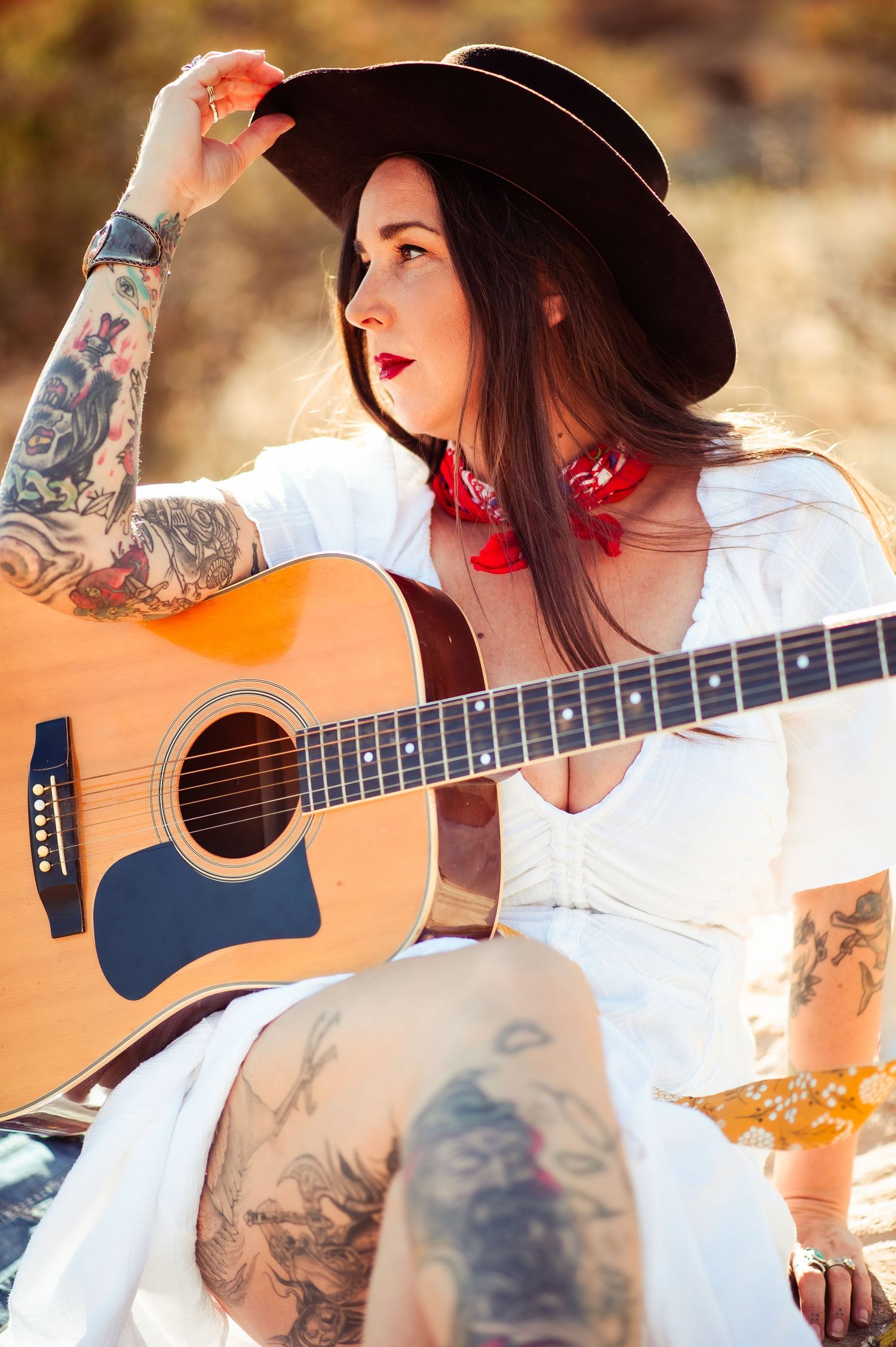 Musician with tattoos playing acoustic guitar while wearing a black hat and white outfit.