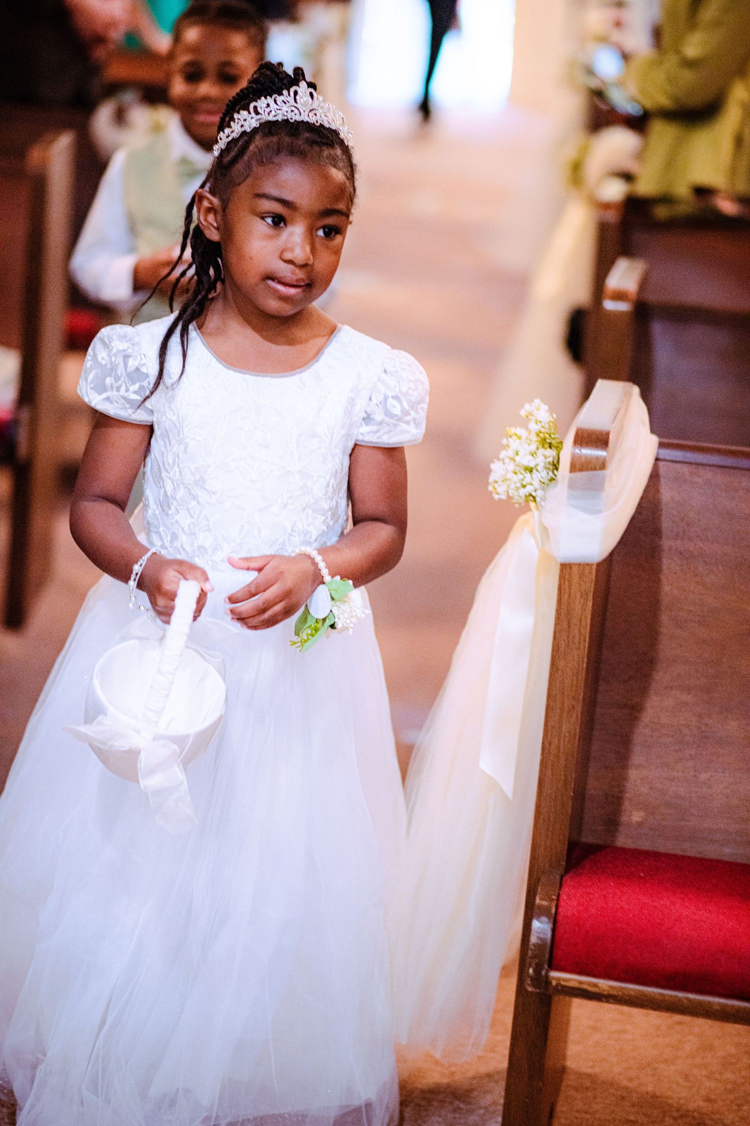 Young flower girl in white lace dress and tiara holds basket while walking down church aisle at wedding ceremony.