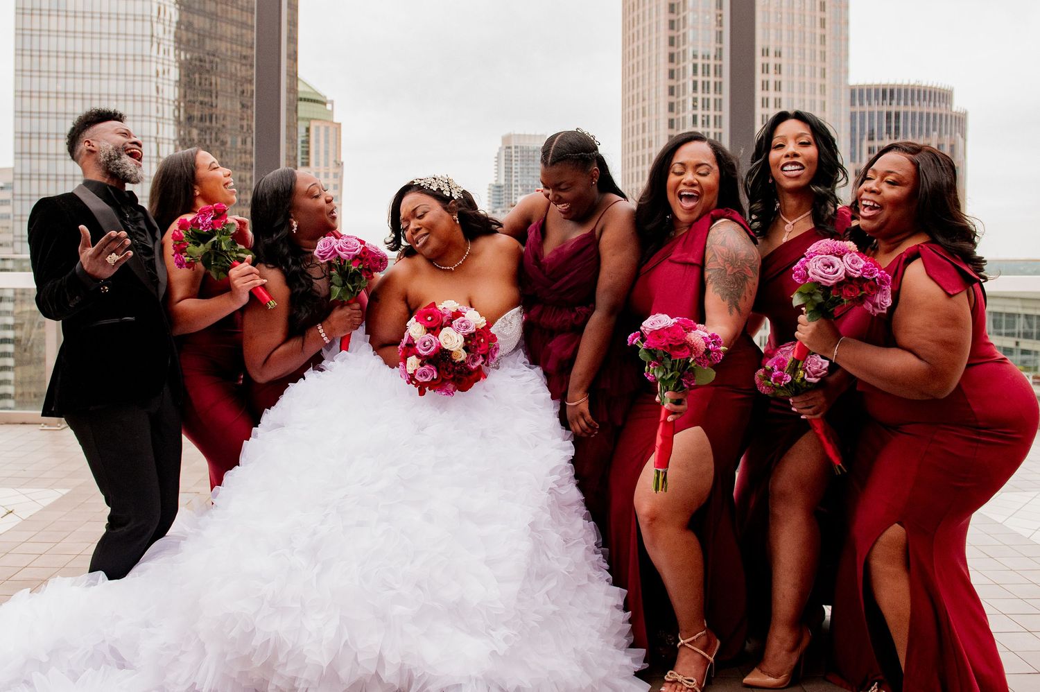 Wedding party in burgundy dresses laughs together during outdoor photo session with bride in white gown.