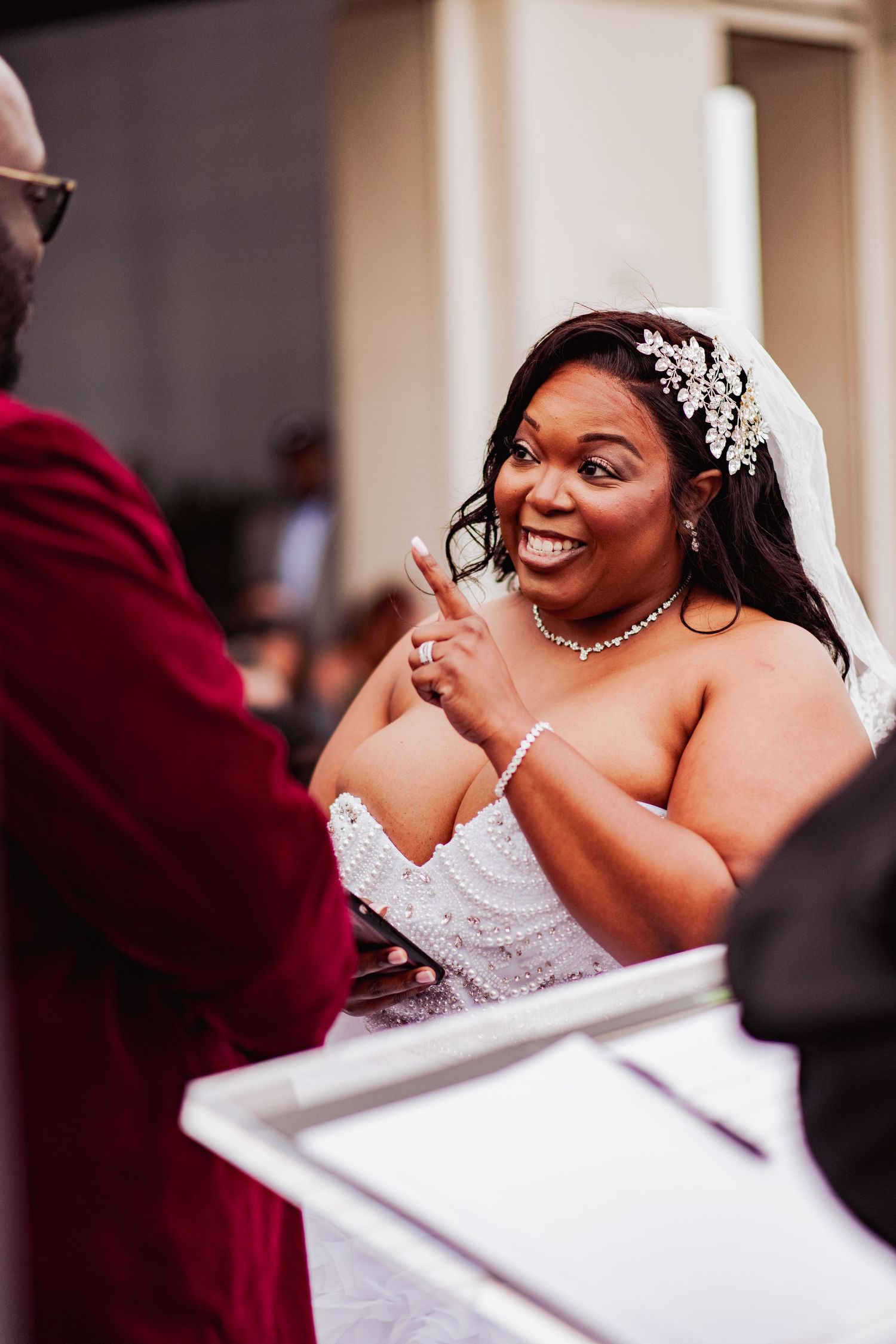 A radiant bride in a white lace gown and crystal headpiece smiles during her wedding ceremony.