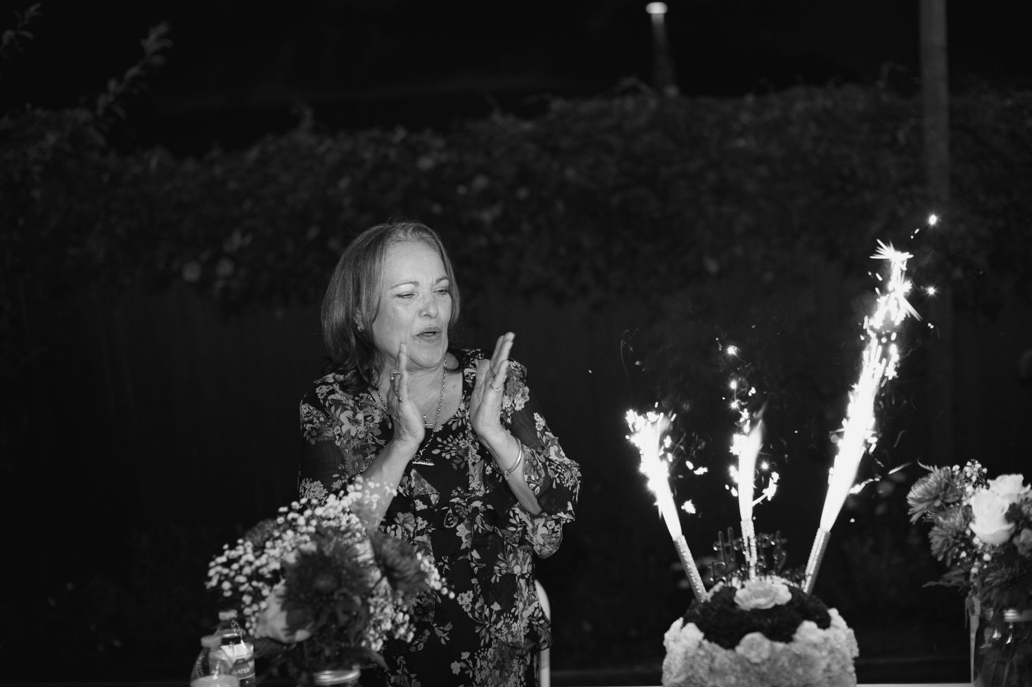 A black and white photo shows someone clapping near a cake with sparkler candles at a nighttime celebration.