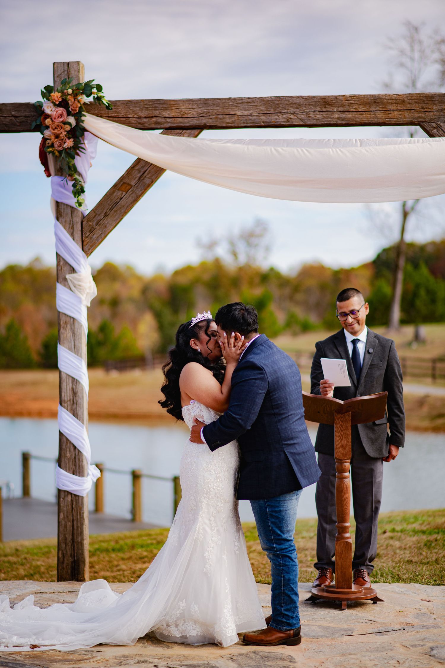 A couple shares a romantic moment under a white canopy by a lake during an outdoor wedding ceremony.