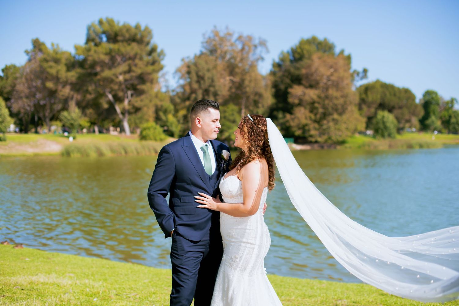 Sunlit wedding portrait by a Whittier Narrows lake with the bride's long veil catching the breeze.