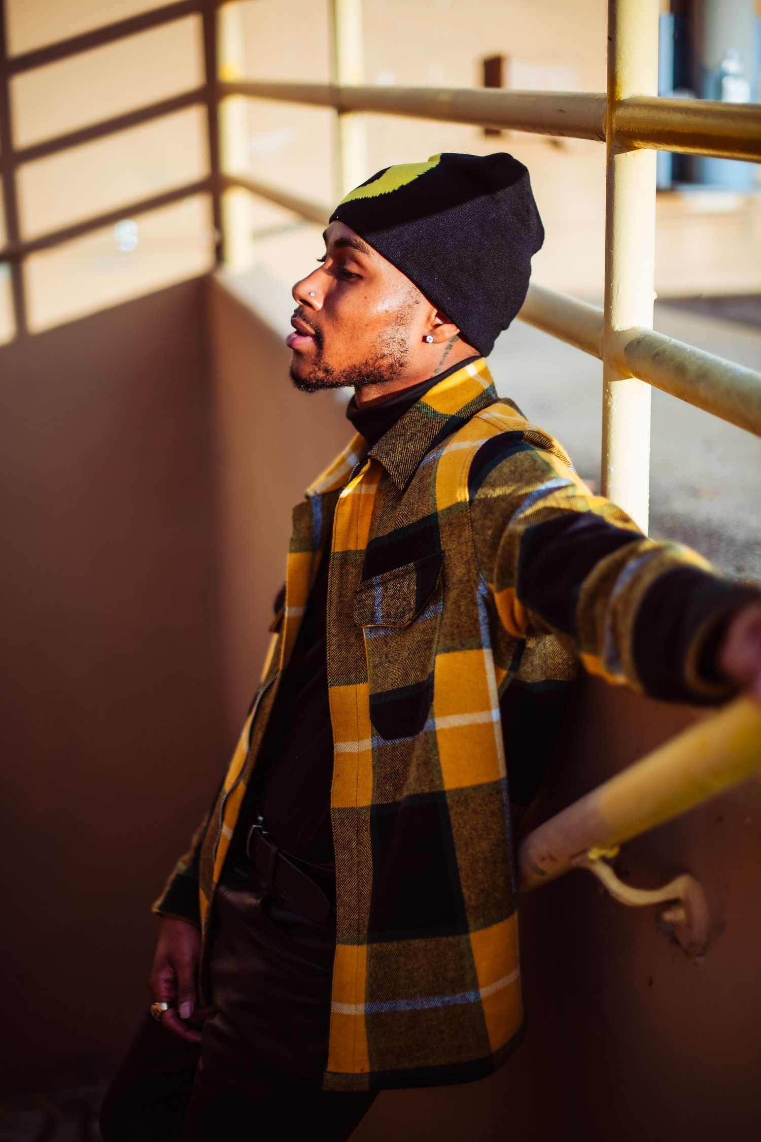 Person in black beanie and yellow plaid flannel jacket poses against wall with dramatic lighting and shadows.