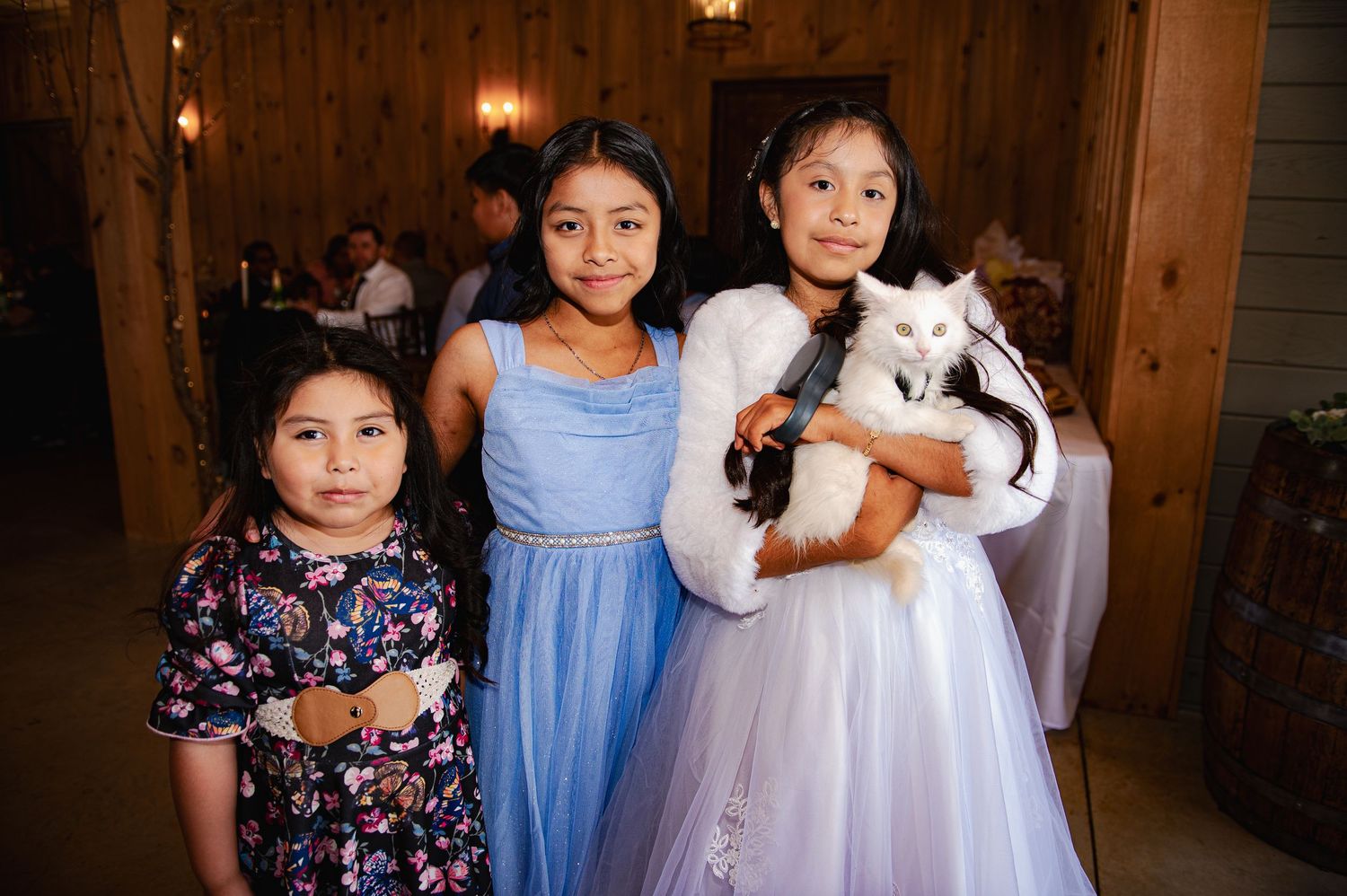 Three young girls in party dresses pose together at an indoor venue, with one holding a white cat.