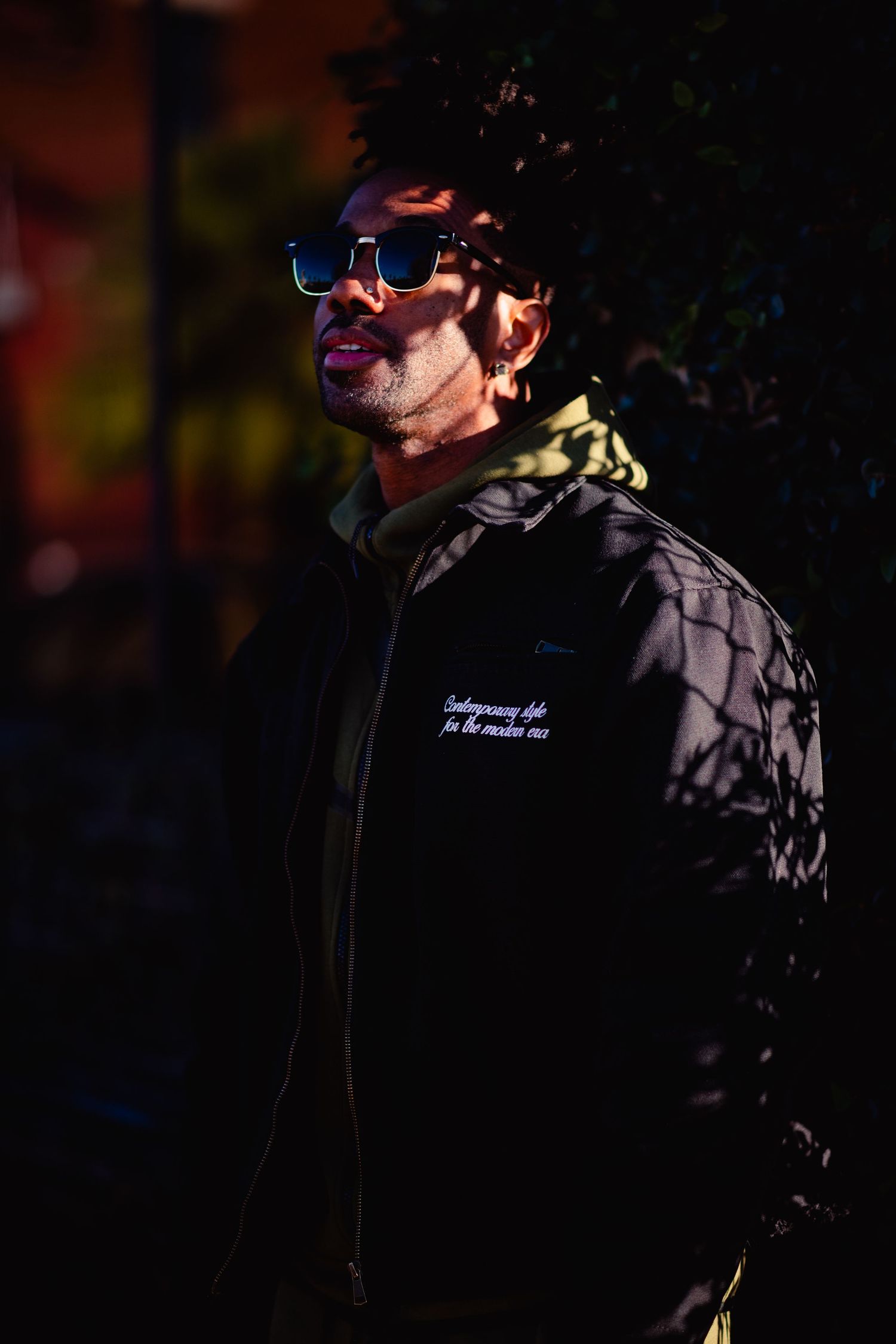 A performer in dark clothing and sunglasses stands against a shadowy background during a stage performance.