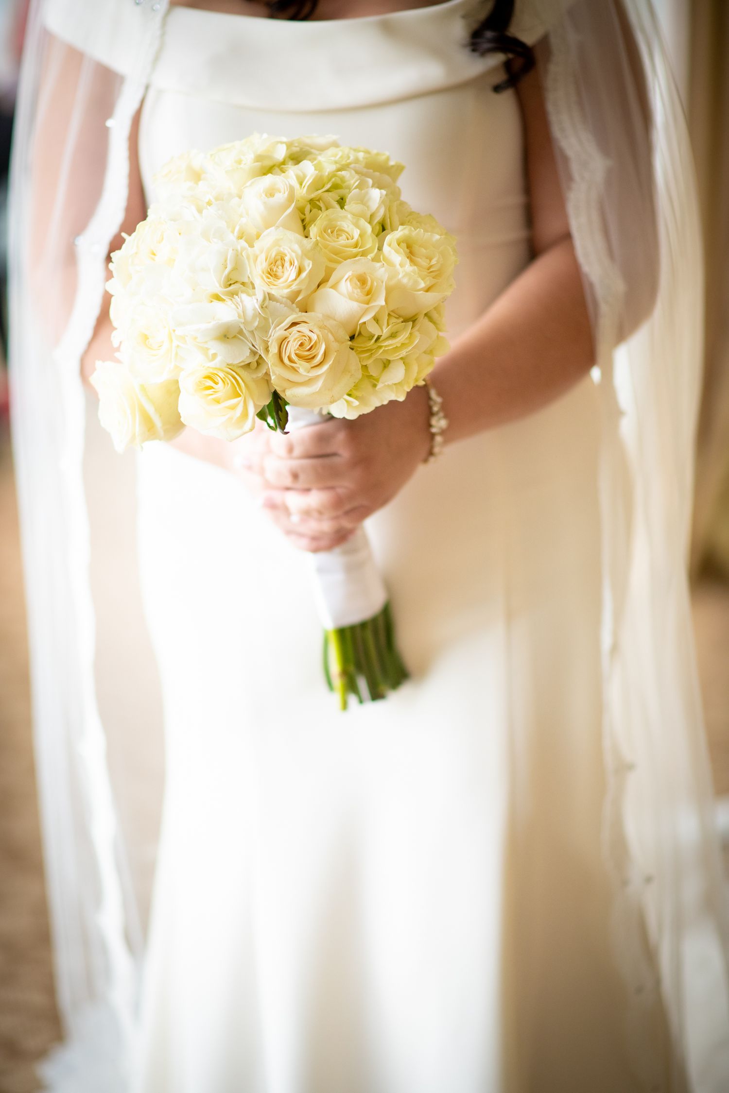 Bride holding an elegant bouquet of cream colored roses against a white wedding dress in soft lighting.