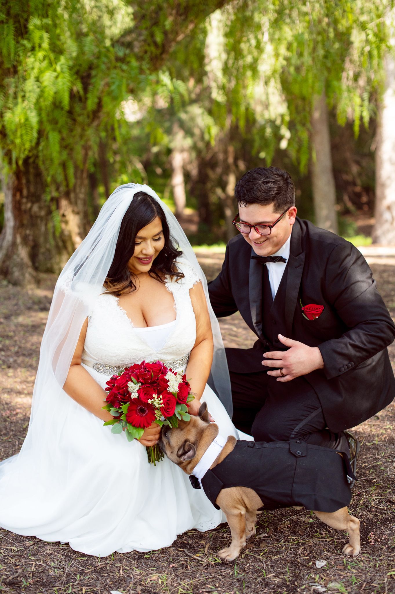 A couple in wedding attire shares a playful moment in a wooded outdoor setting with red roses in the bouquet.
