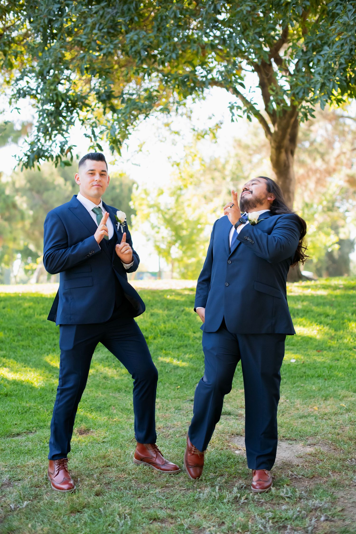 Two groomsmen in navy suits strike playful poses on grass under a tree.