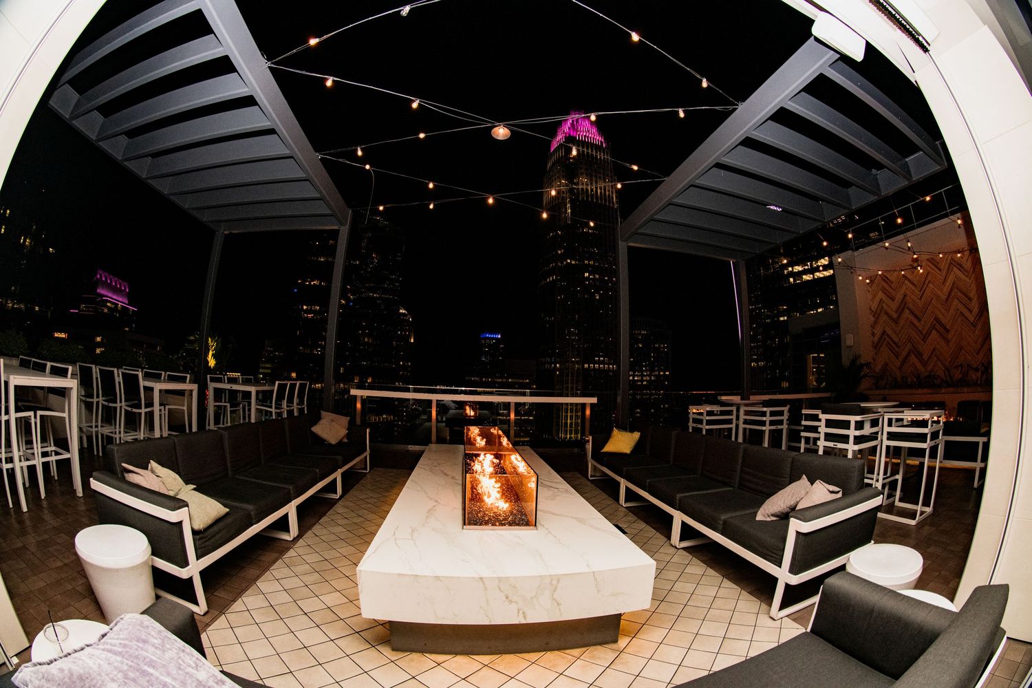 Modern rooftop lounge setup with fire pit and string lights creating an intimate outdoor atmosphere.
