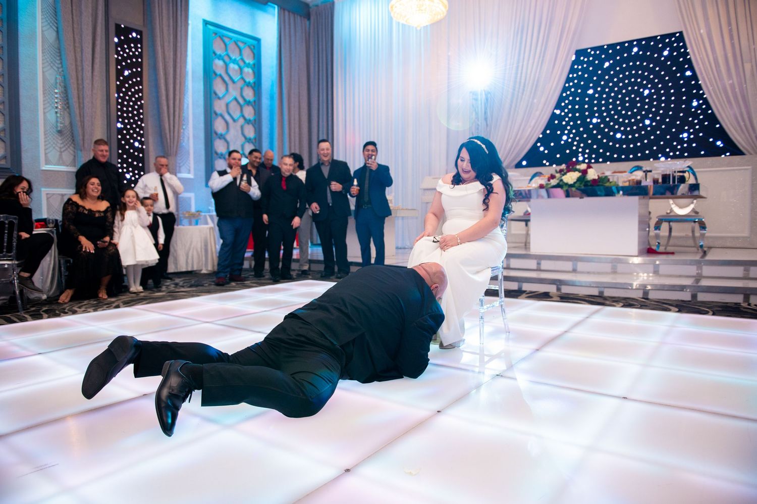 Wedding guests dance and jump on an illuminated white dance floor at an elegant reception venue.