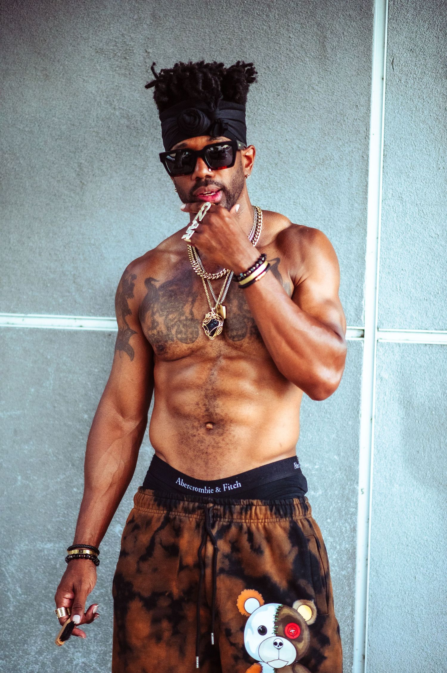 Shirtless muscular man in tie-dye shorts with cartoon bear design poses against white wall.