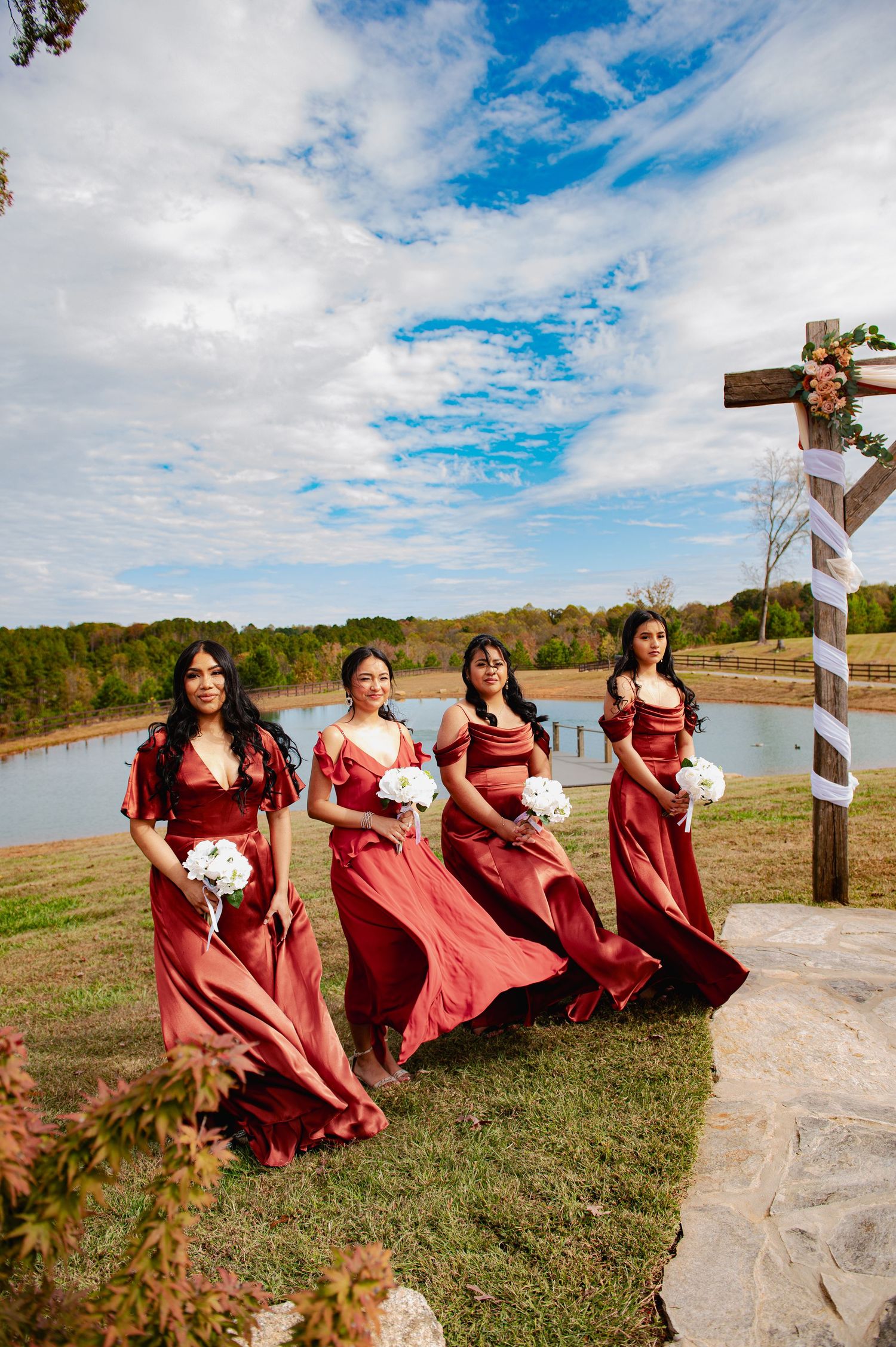 Bridesmaids in burgundy dresses pose playfully by a lake on a cloudy autumn day.