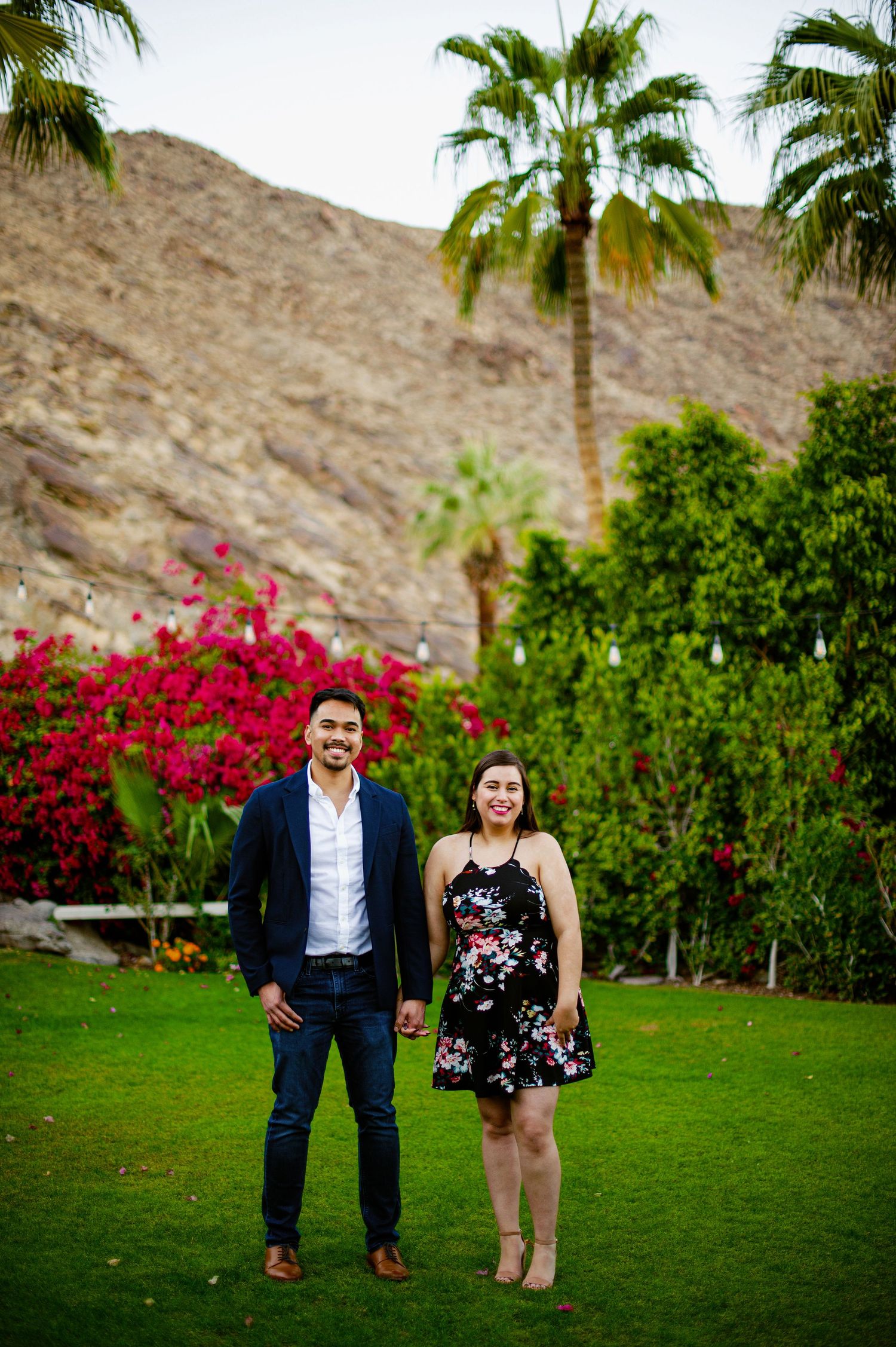 A couple poses together on a lush green lawn with palm trees and vibrant pink bougainvillea flowers in the desert landscape.