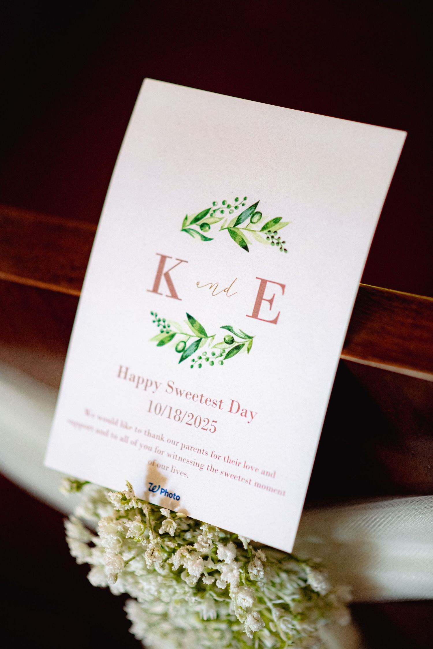 Wedding invitation cards with floral designs and monogram initials displayed with white flowers on a dark wooden surface.