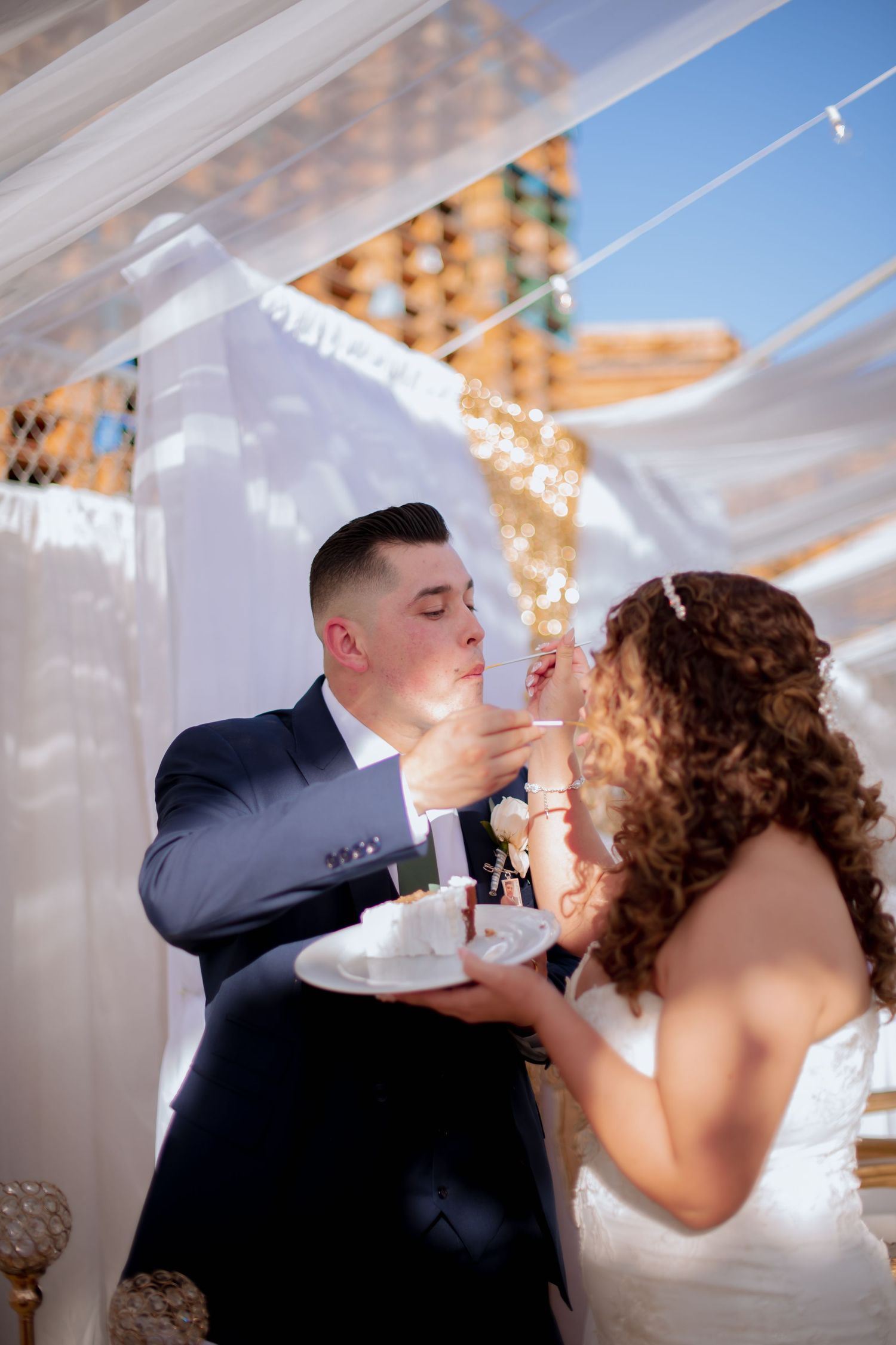 Newlyweds sharing wedding cake during their reception under white draped ceiling.