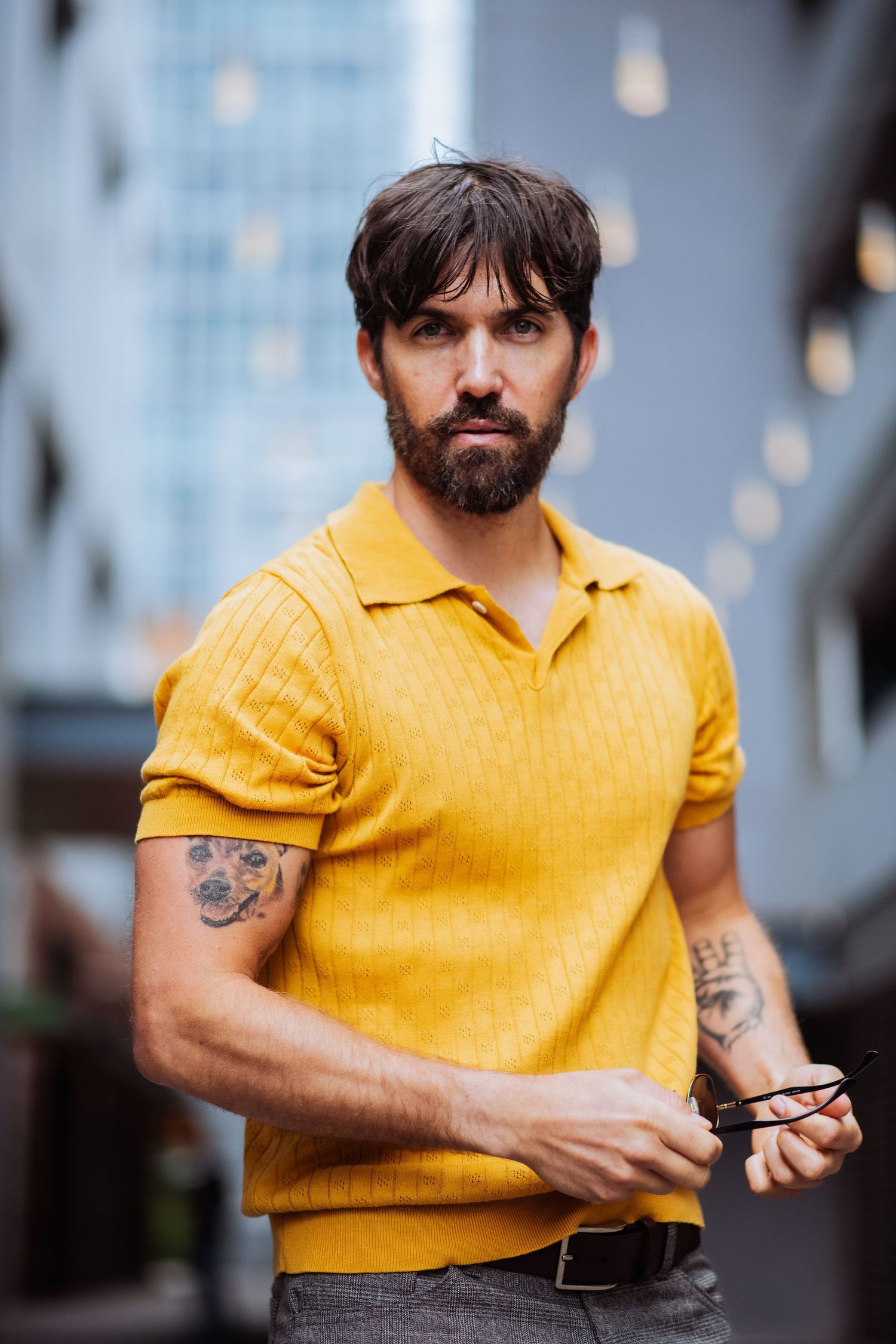 Person with tattoos wearing yellow polo checks watch while standing in urban setting.