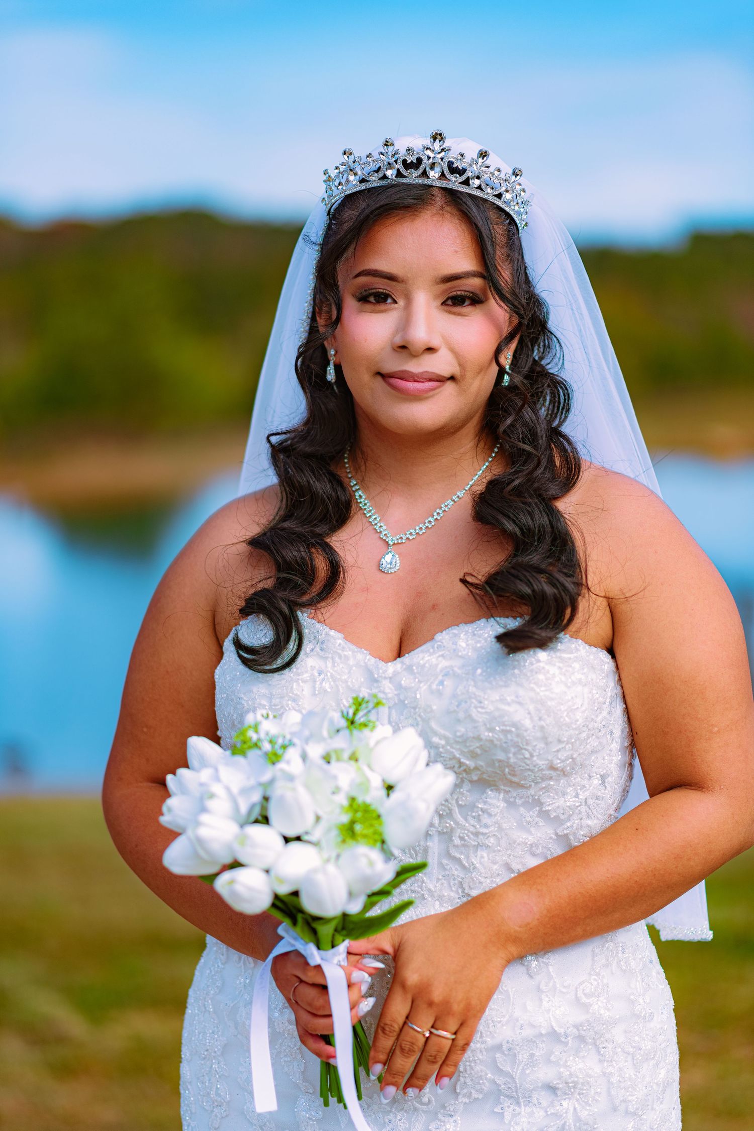 Elegant bride in a white lace dress and tiara holds a white floral bouquet against a lakeside backdrop.