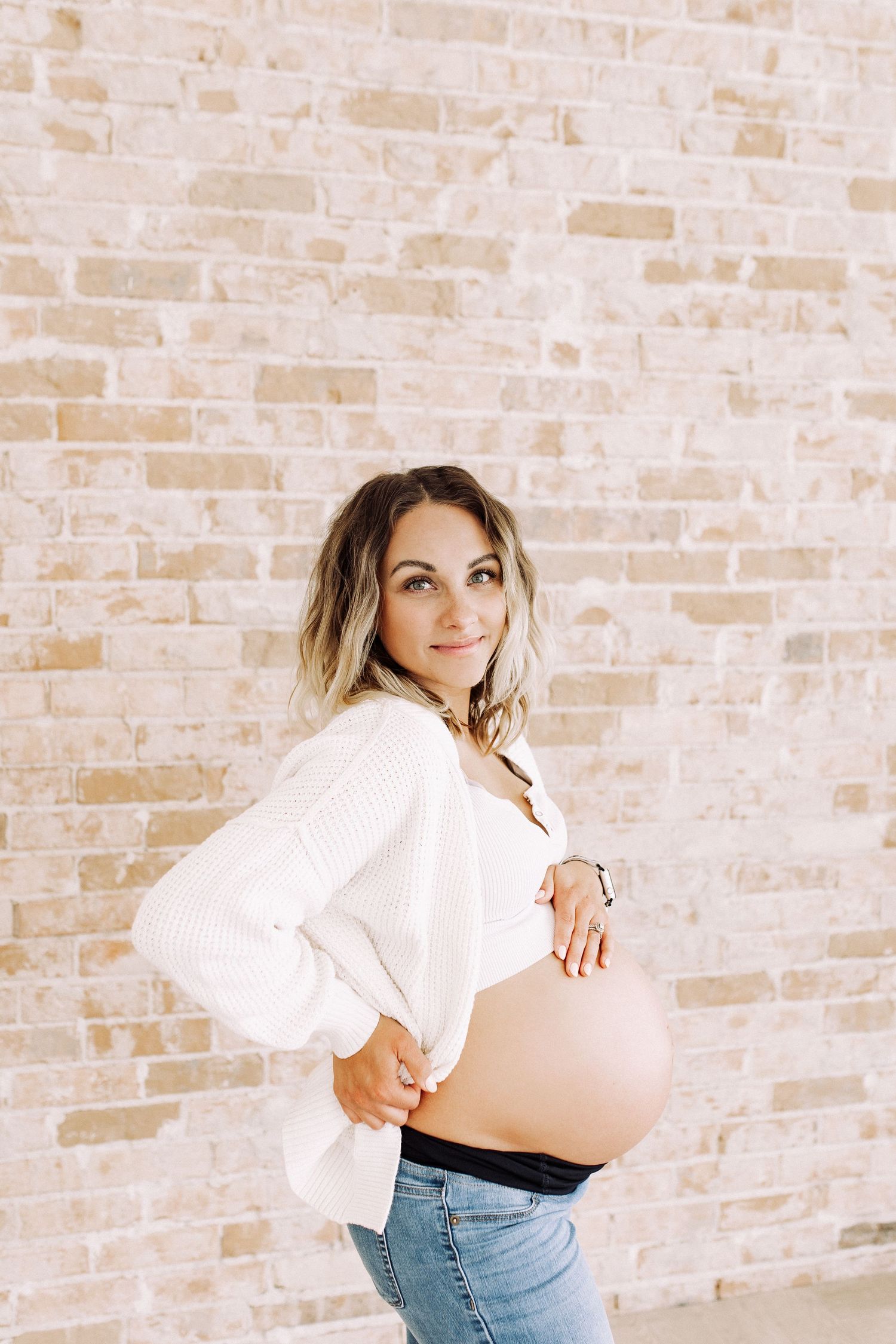 Maternity portraits of woman in white shirt and jeans against brick wall.