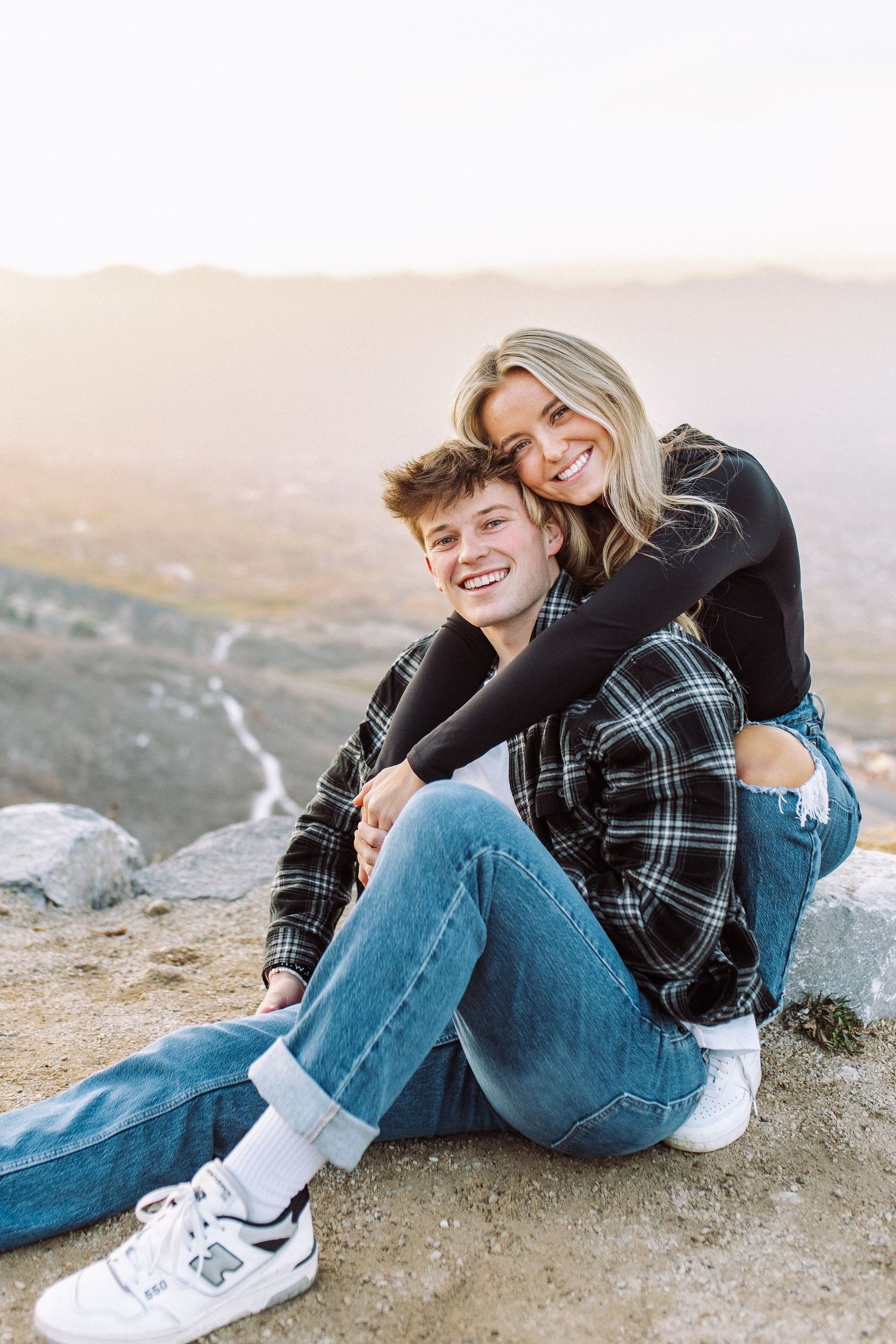 A couple in casual wear sits together on a rocky overlook during sunset.