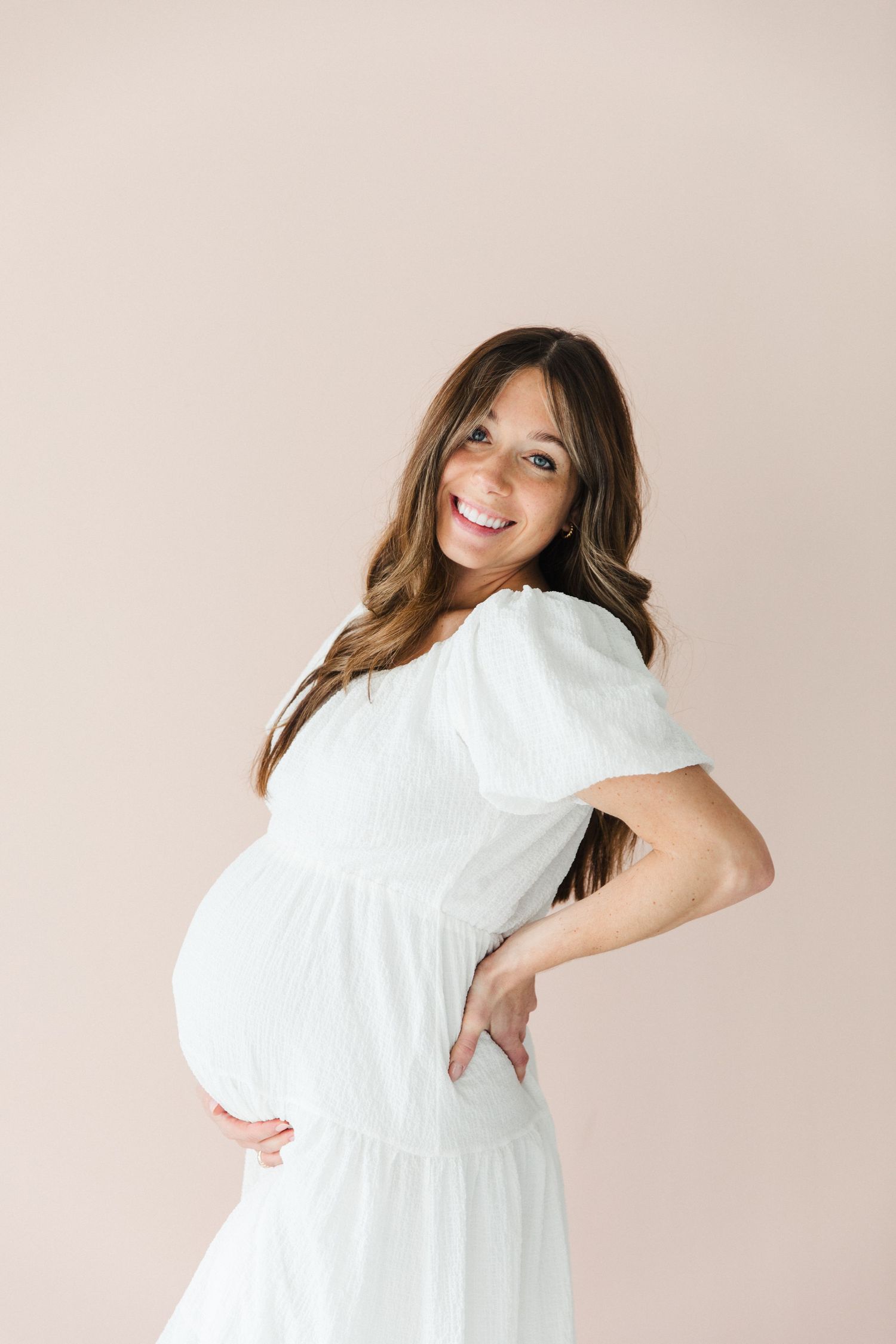 Expectant mother in white dress poses sideways showing her baby bump against neutral background.
