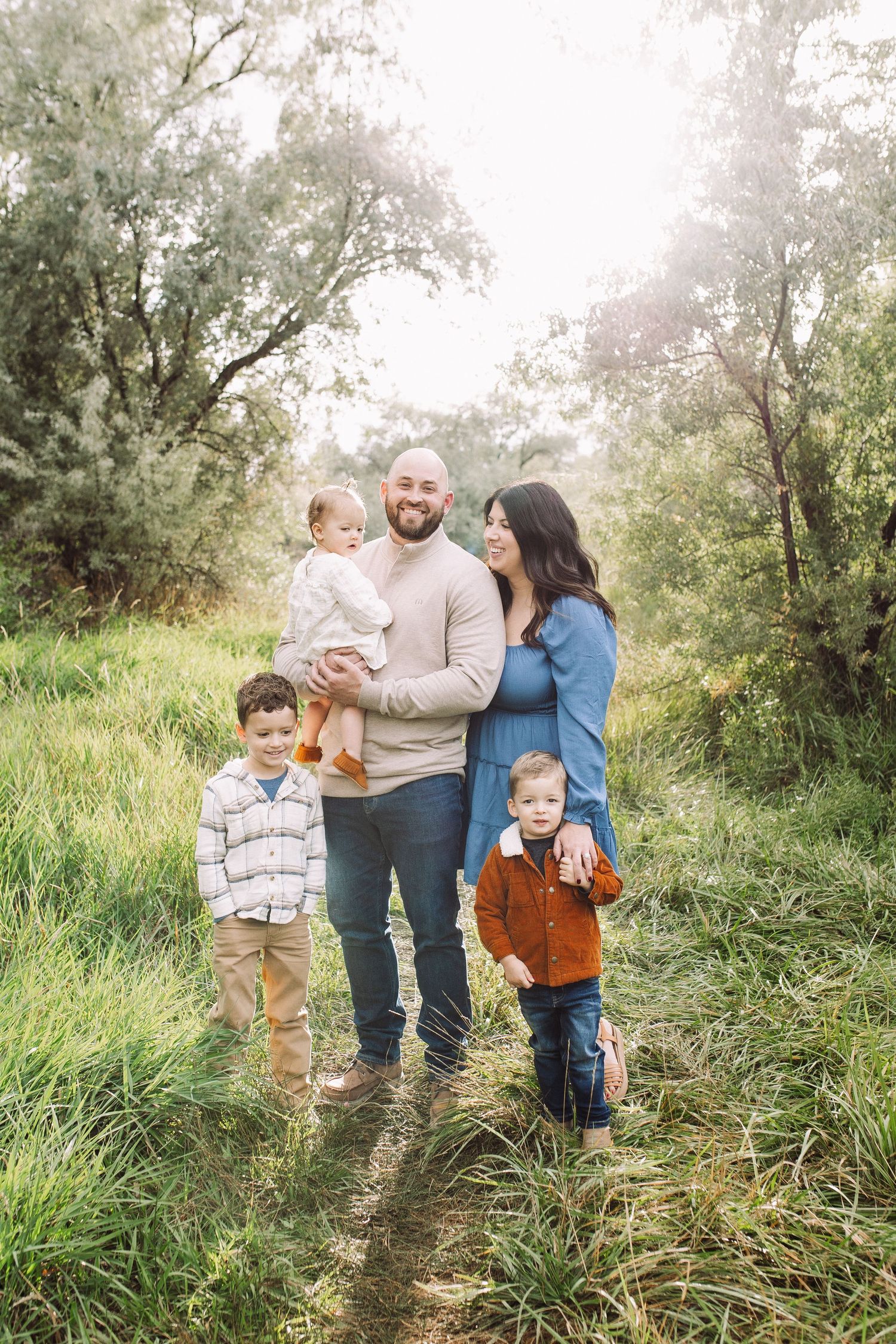 Family candid moment captured in natural setting with tall grass and sunlit trees.